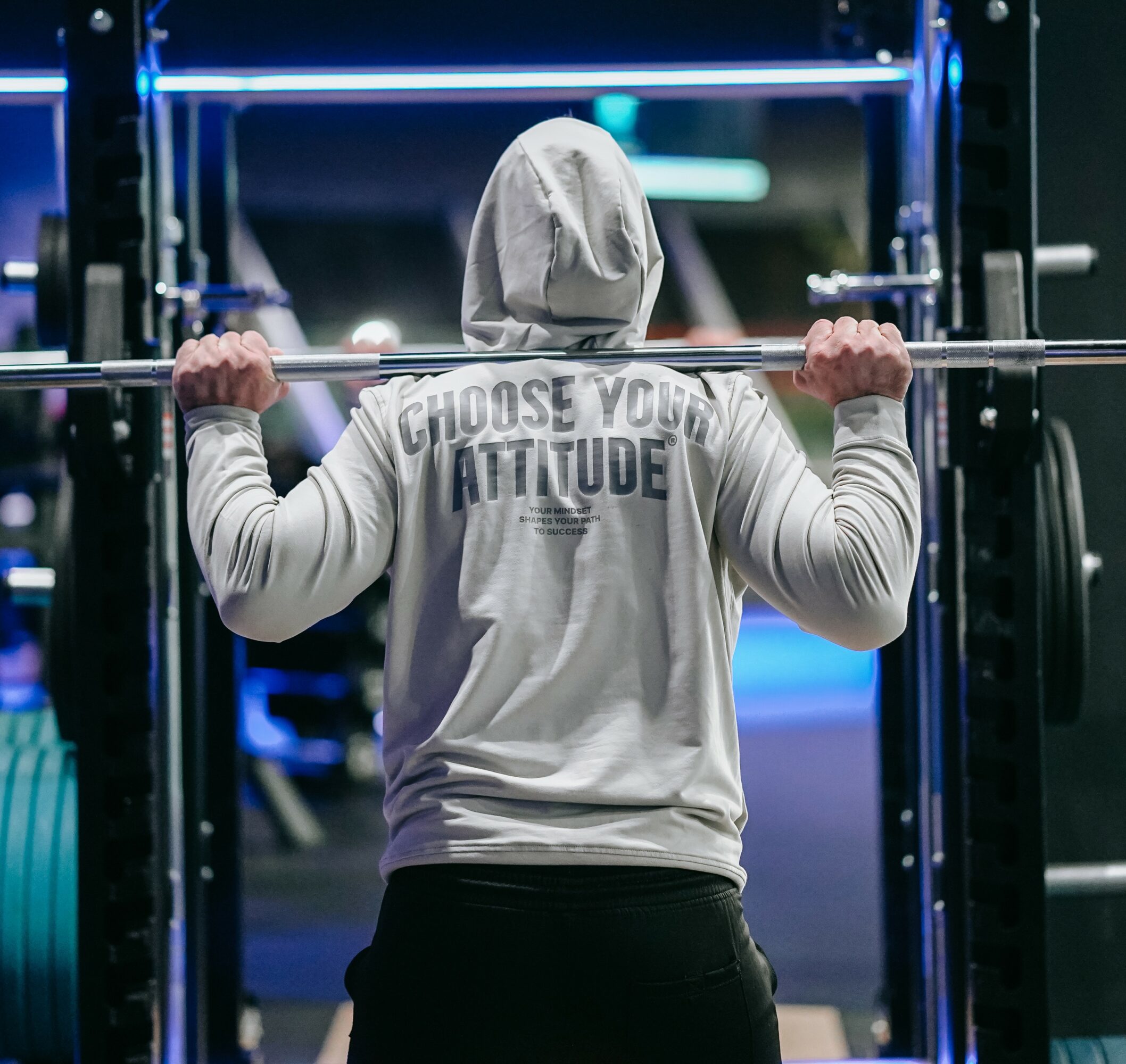 Person lifting a barbell in a gym, wearing a hoodie with the text 'CHOOSE YOUR ATTITUDE' on the back.