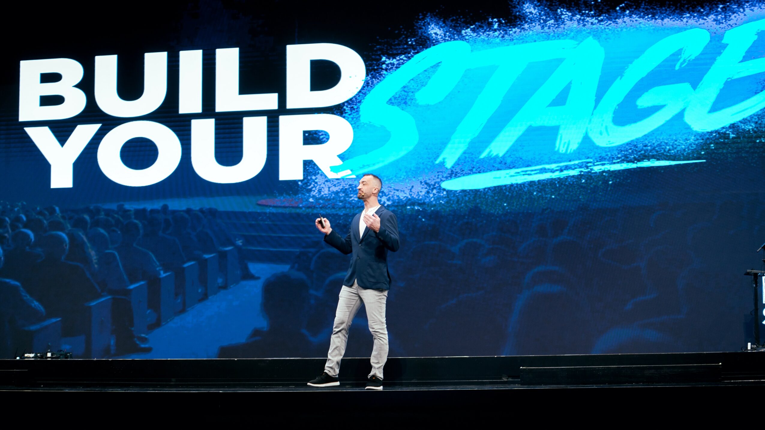 Person standing on stage in front of a large screen with the text 'BUILD YOUR STAGE' in bold letters.