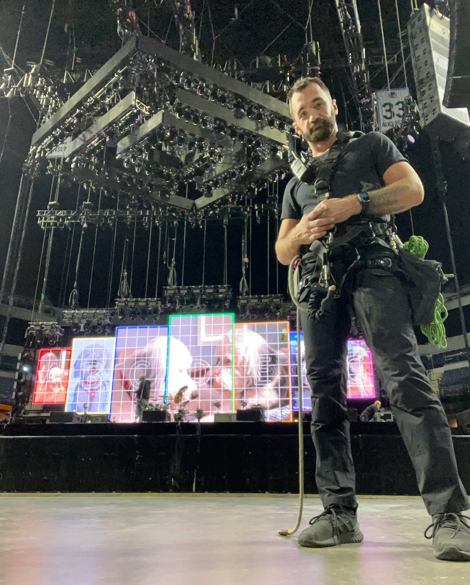 Man standing on stage with lighting and equipment overhead, holding a camera, in a concert or event venue.