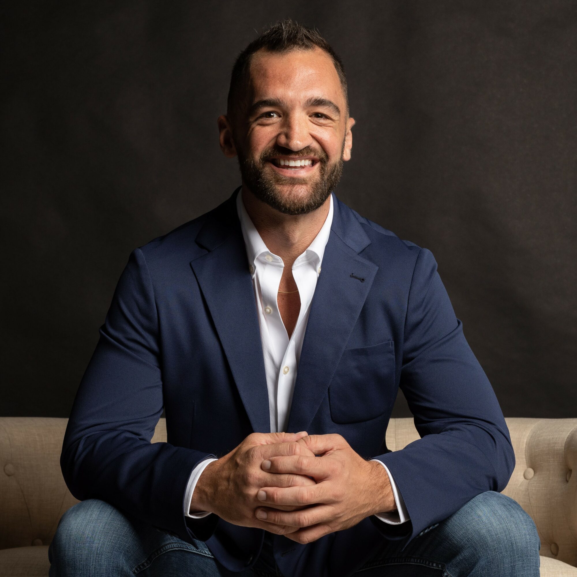 Man with a beard smiling, wearing a navy blazer and white shirt, sitting with hands clasped, dark background.