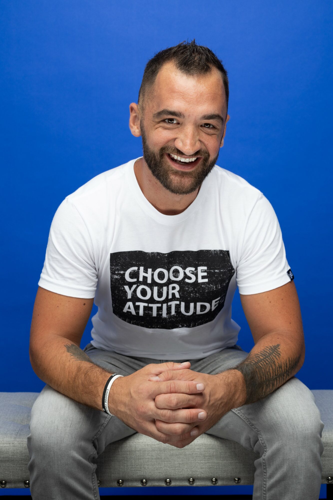 Smiling man with a beard and short hair, wearing a white T-shirt with text, sitting against a blue background.