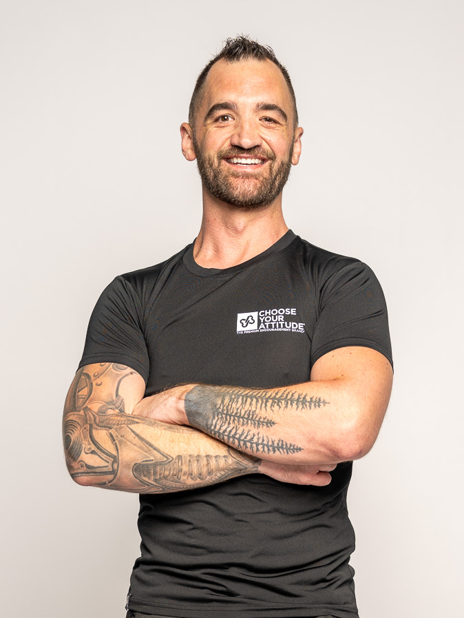 Smiling man with tattoos on arms, wearing a black t-shirt, arms crossed, against a plain background.