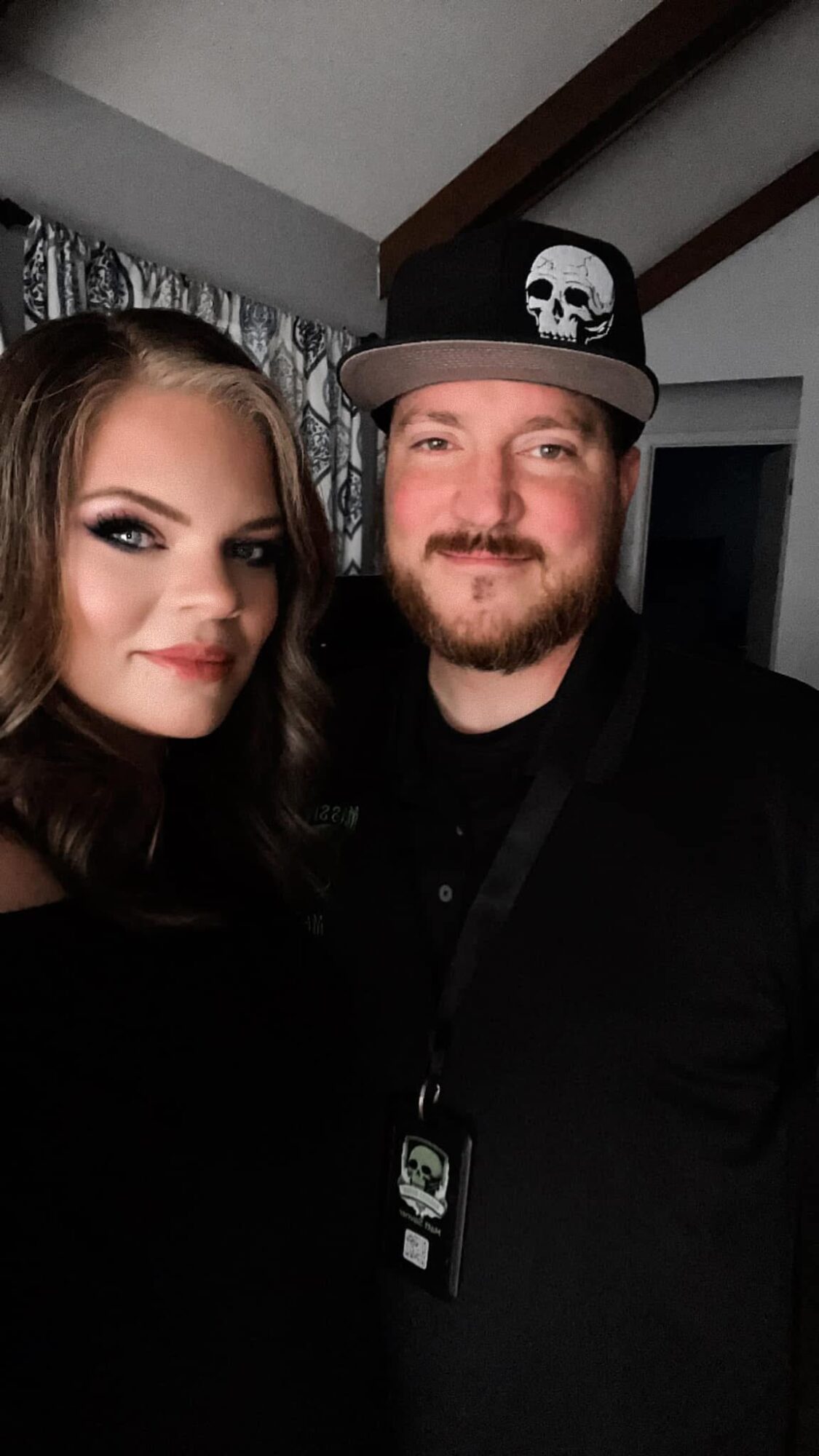 Two people posing indoors, one wearing a cap with a skull, both smiling, dark clothing, background with curtains and ceiling beams.