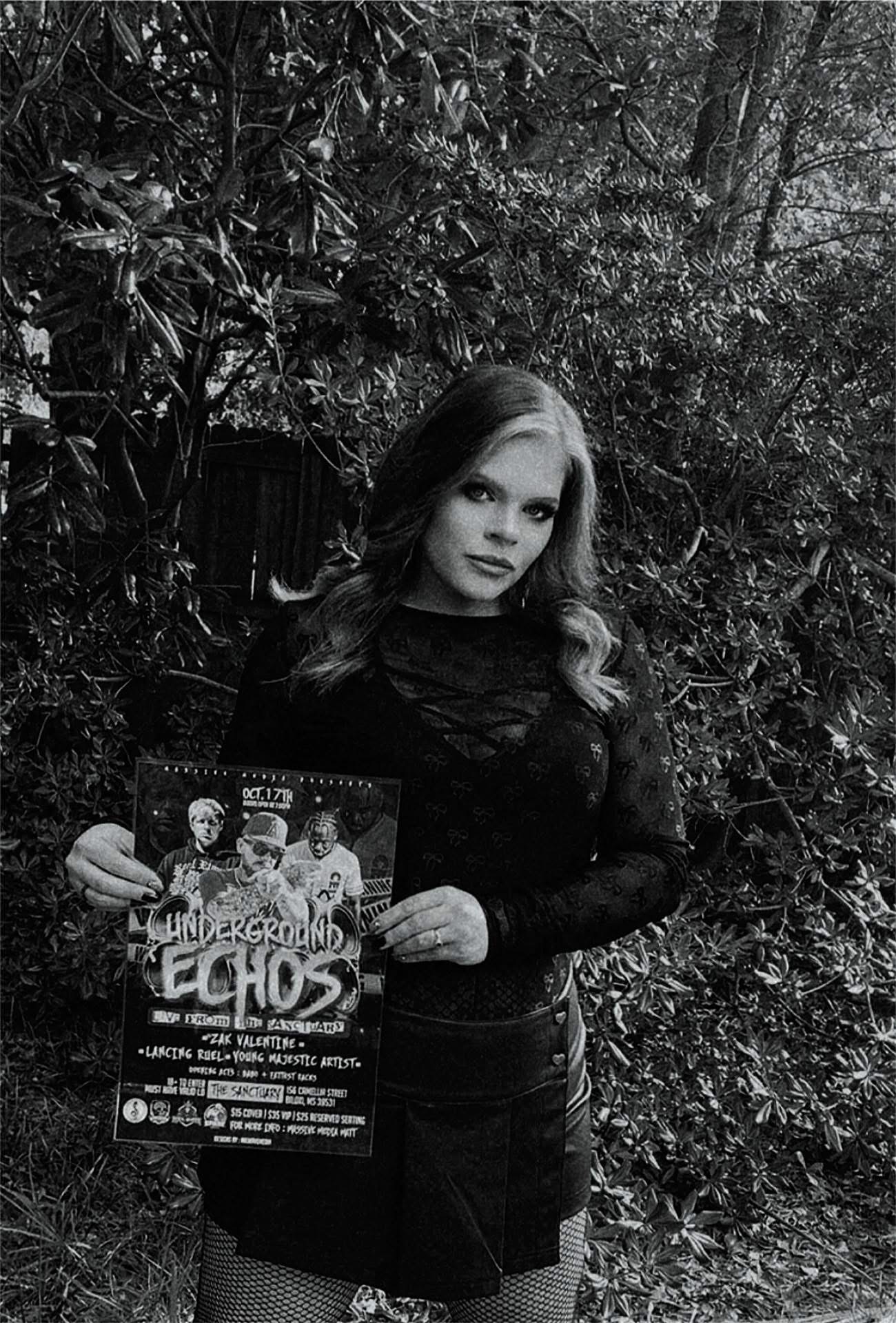 Young woman with wavy hair holding a poster outdoors, surrounded by trees and foliage, in black and white.