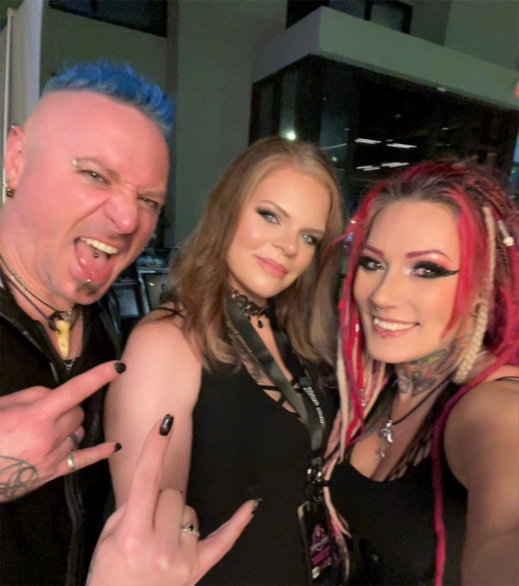 Three people smiling and posing for a photo indoors, with colorful hair and makeup, making rock hand gestures.
