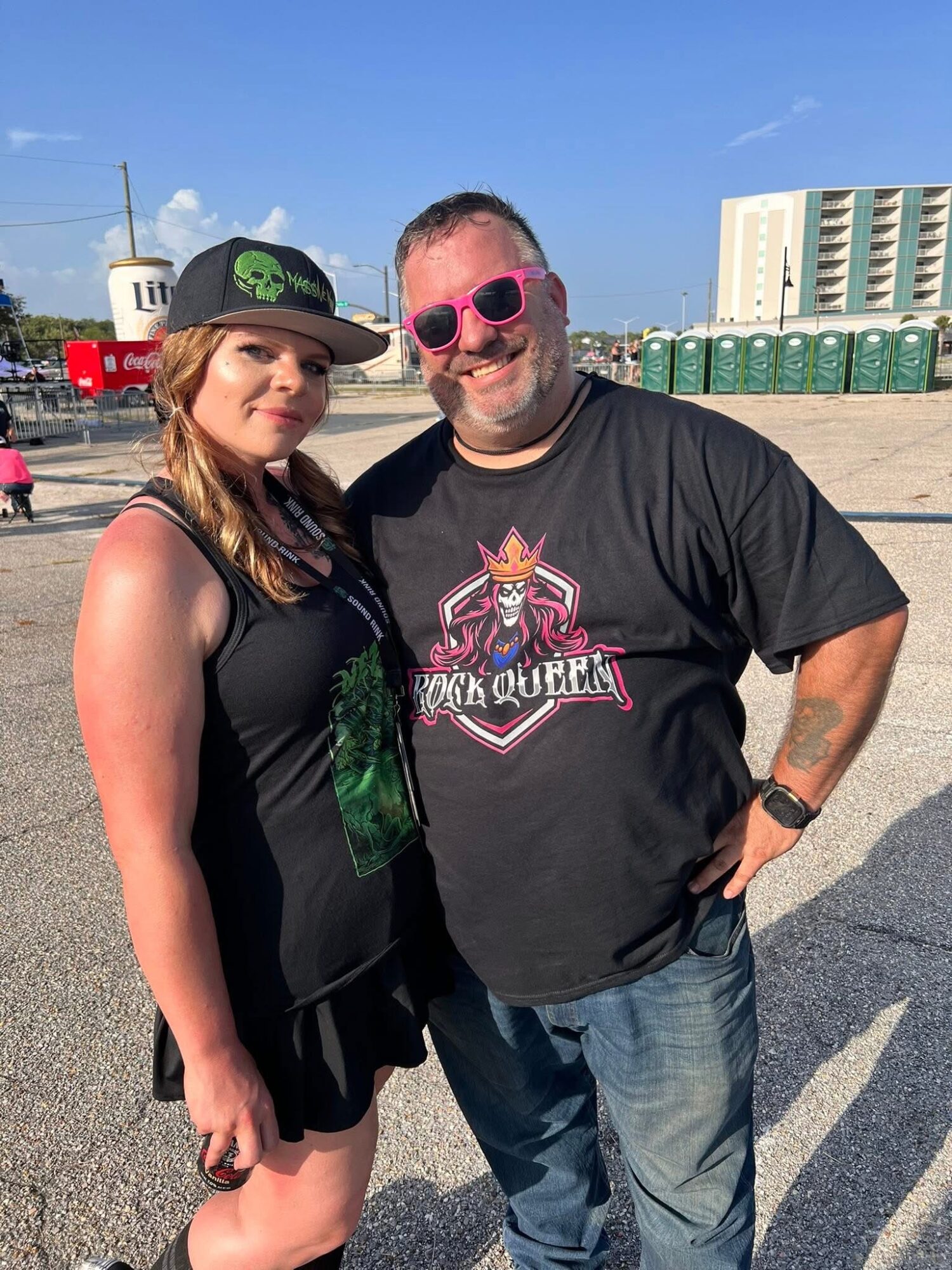 A woman and a man stand outdoors on a sunny day, smiling at the camera, with buildings and a clear sky in background.