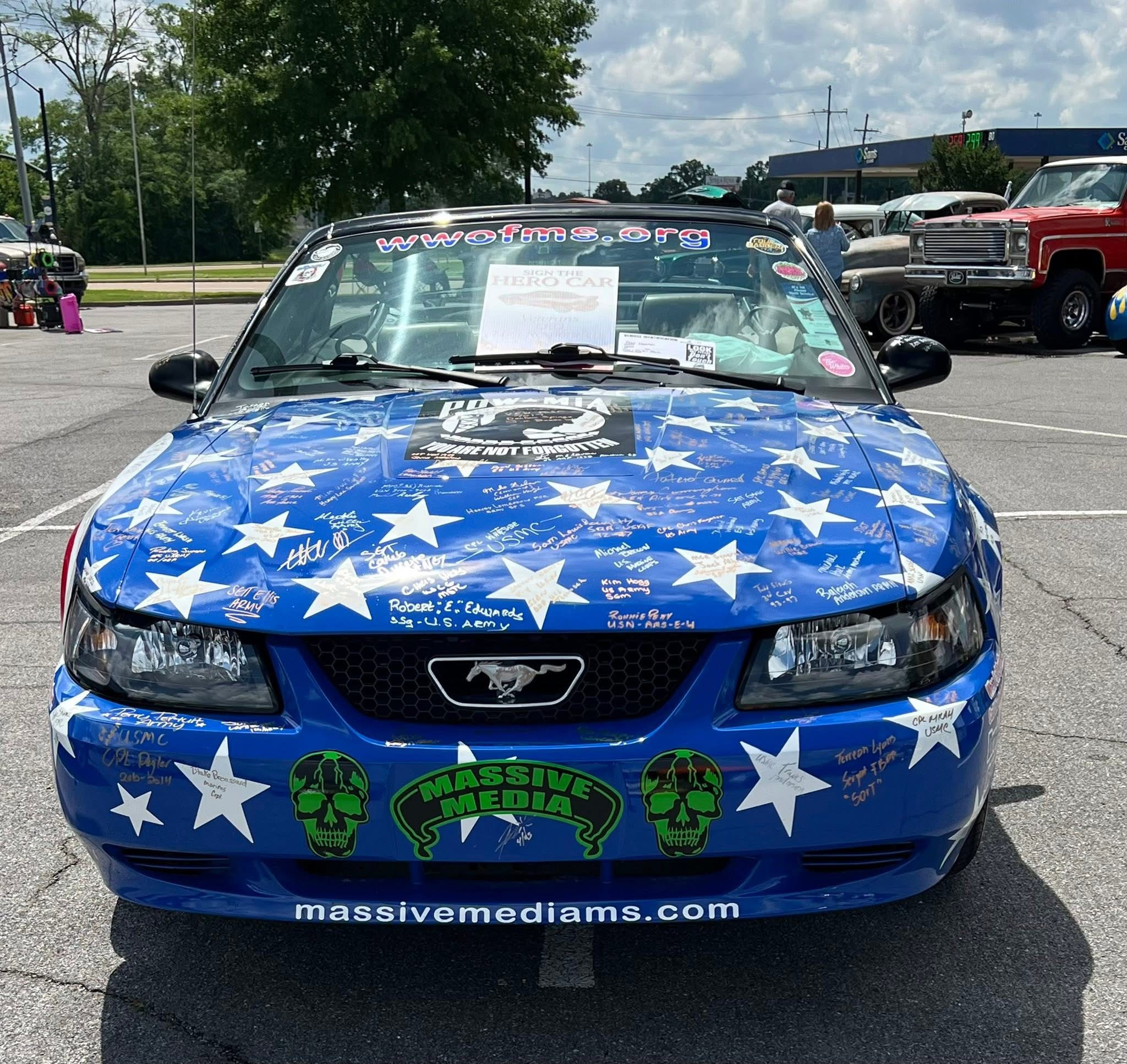 Front view of a blue car with white stars, green skull logos, and website URL, parked outdoors on a sunny day.