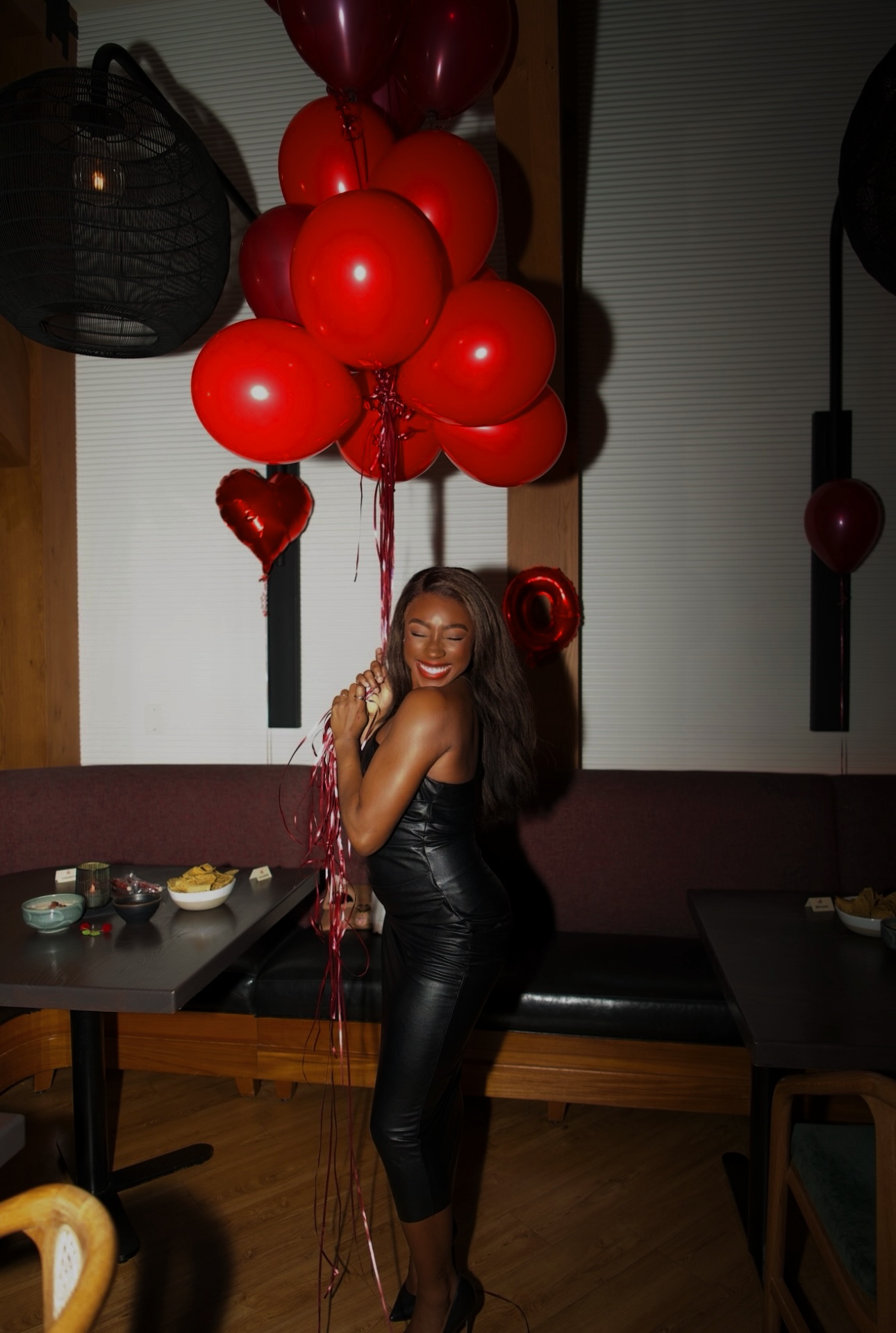 Woman with long hair in black outfit holding red heart-shaped balloons, standing indoors with tables and wall decorations.