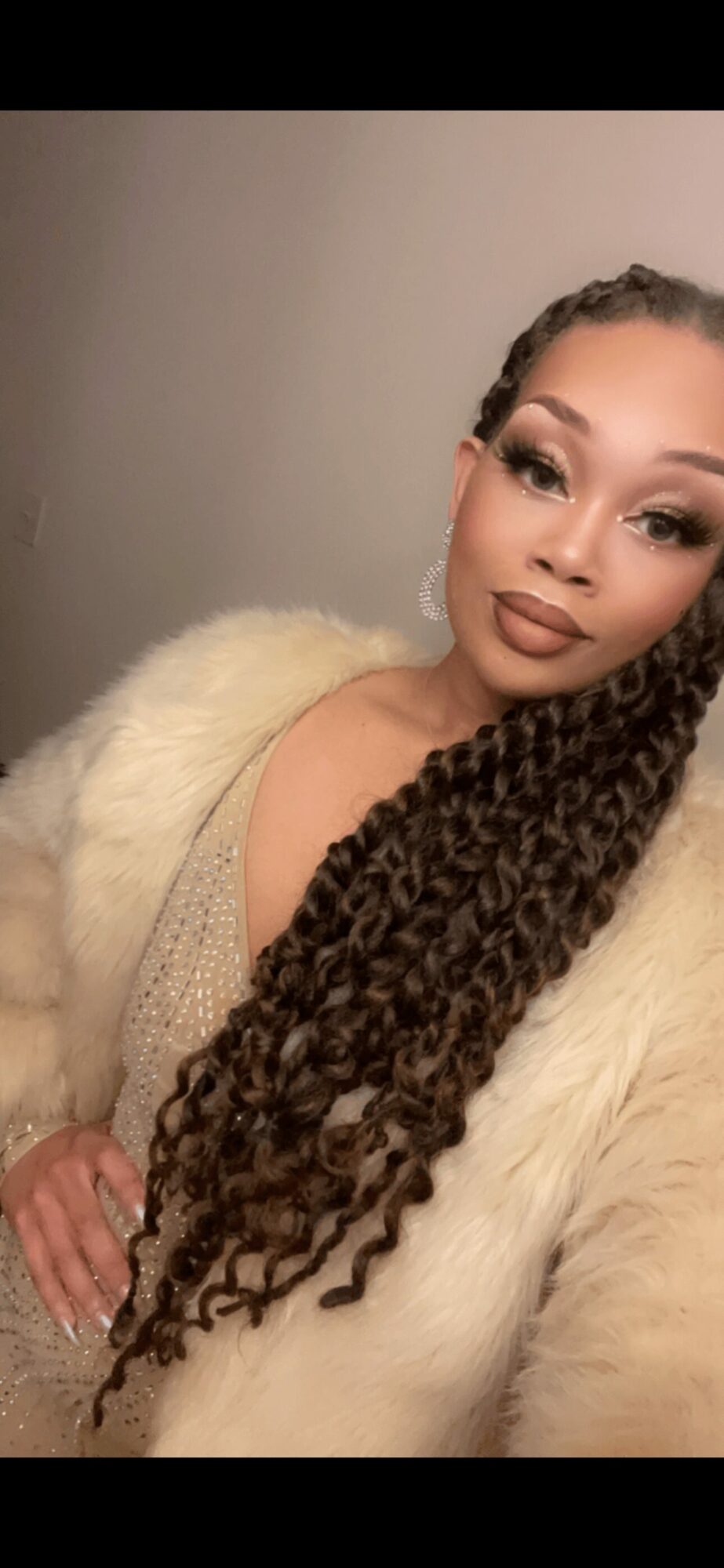 Woman with long braided hair, makeup, and a fur coat, posing indoors against a plain wall.