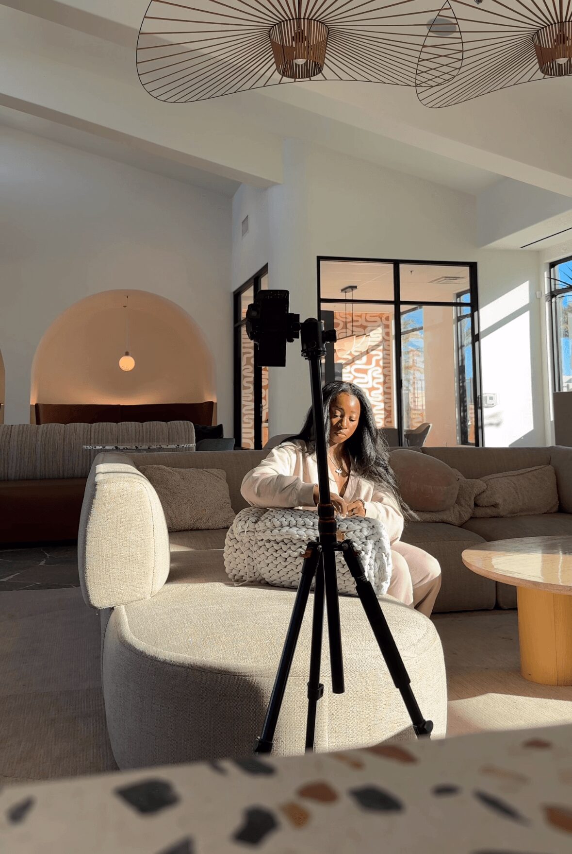 Woman sitting on a sofa in a modern living room, with a tripod in front of her, sunlight streaming in.