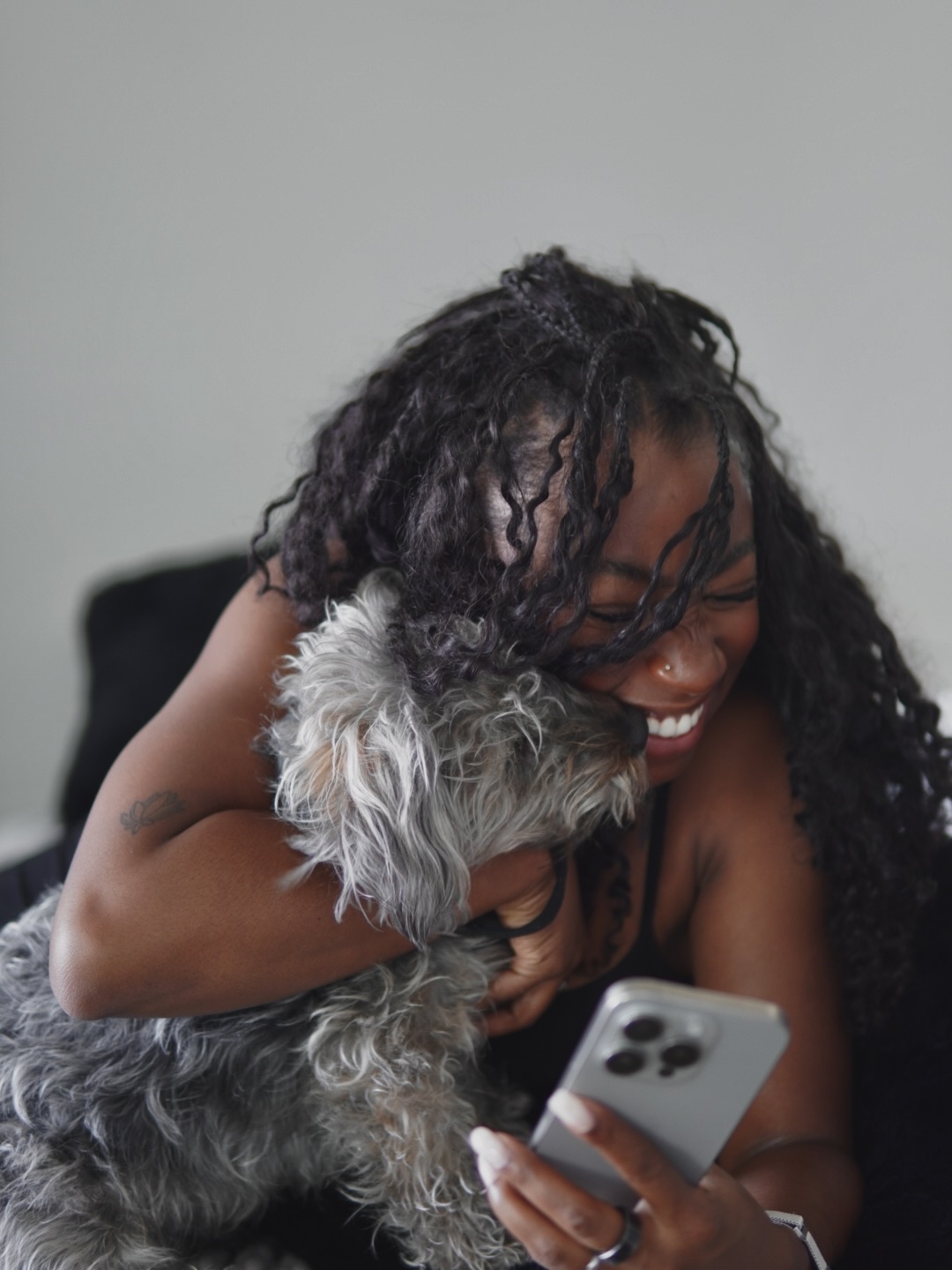 Person with curly hair smiling while holding a small dog and looking at a smartphone.