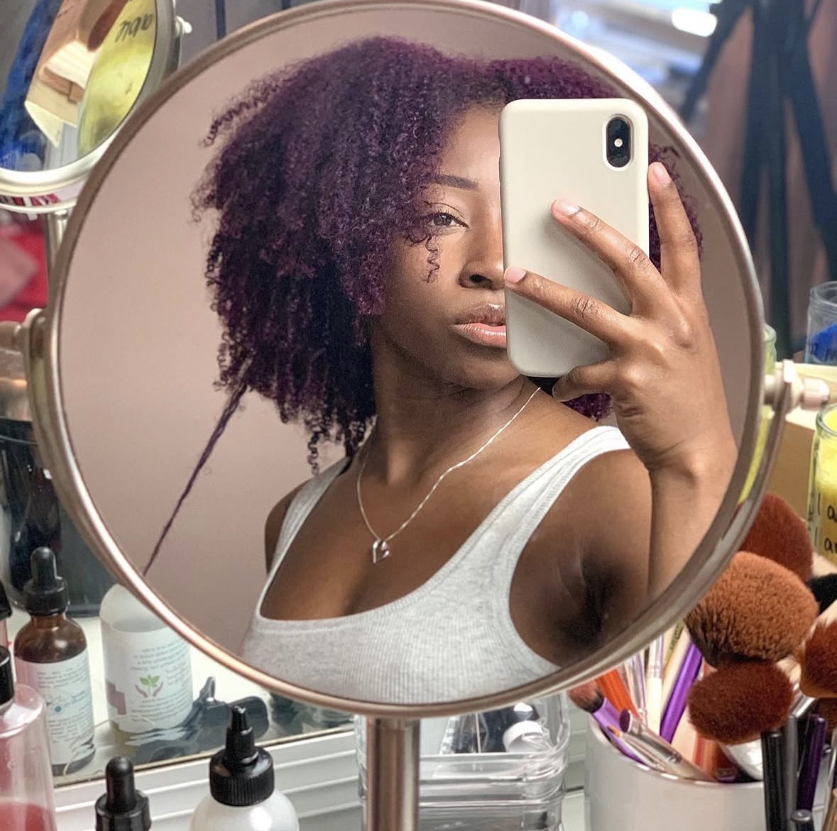 Woman with curly hair taking a mirror selfie, holding a phone, in a room with beauty products.