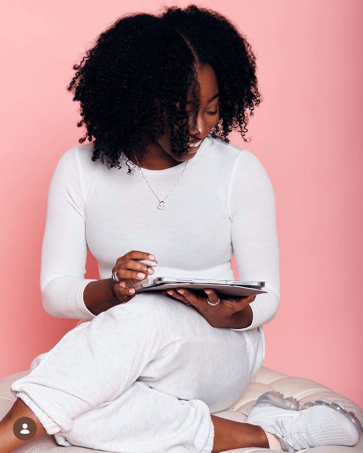 Woman with curly hair sitting cross-legged, looking at a tablet, against a pink background.