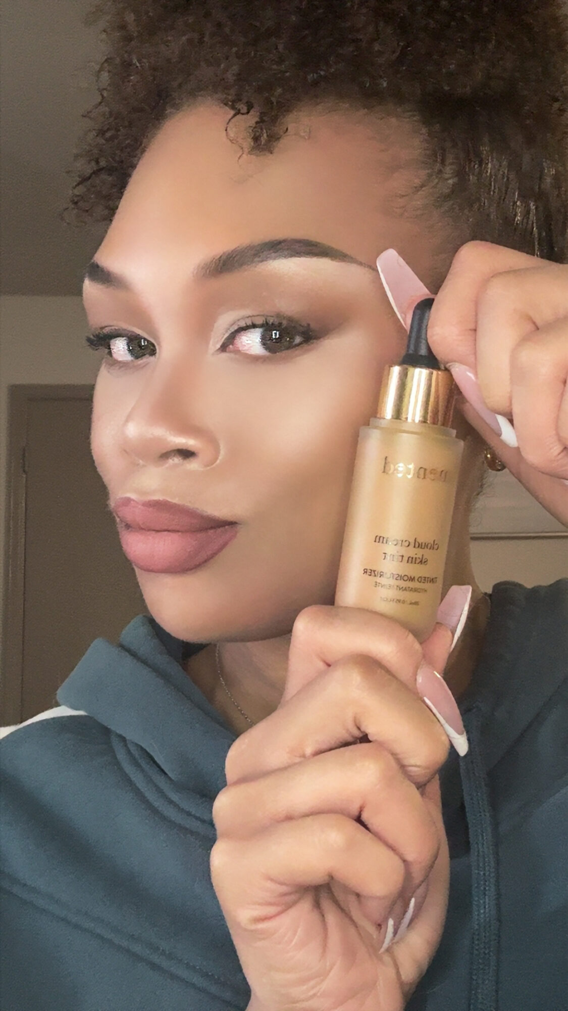 Woman with styled hair holding a gold bottle of skincare product close to her face, smiling softly.