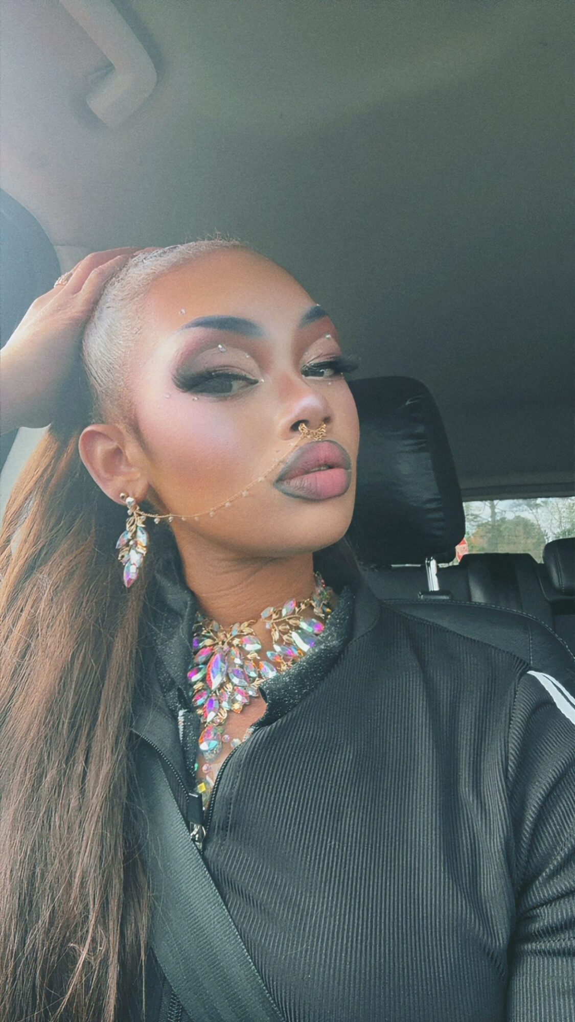 Woman with makeup and jewelry sitting in a car, head tilted, hand on her hair, wearing a black jacket.