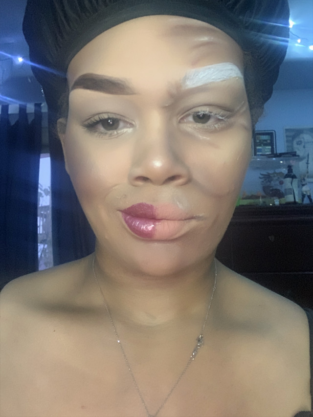 Close-up of a woman with makeup, wearing a black head covering, in a room with shelves and a window behind her.