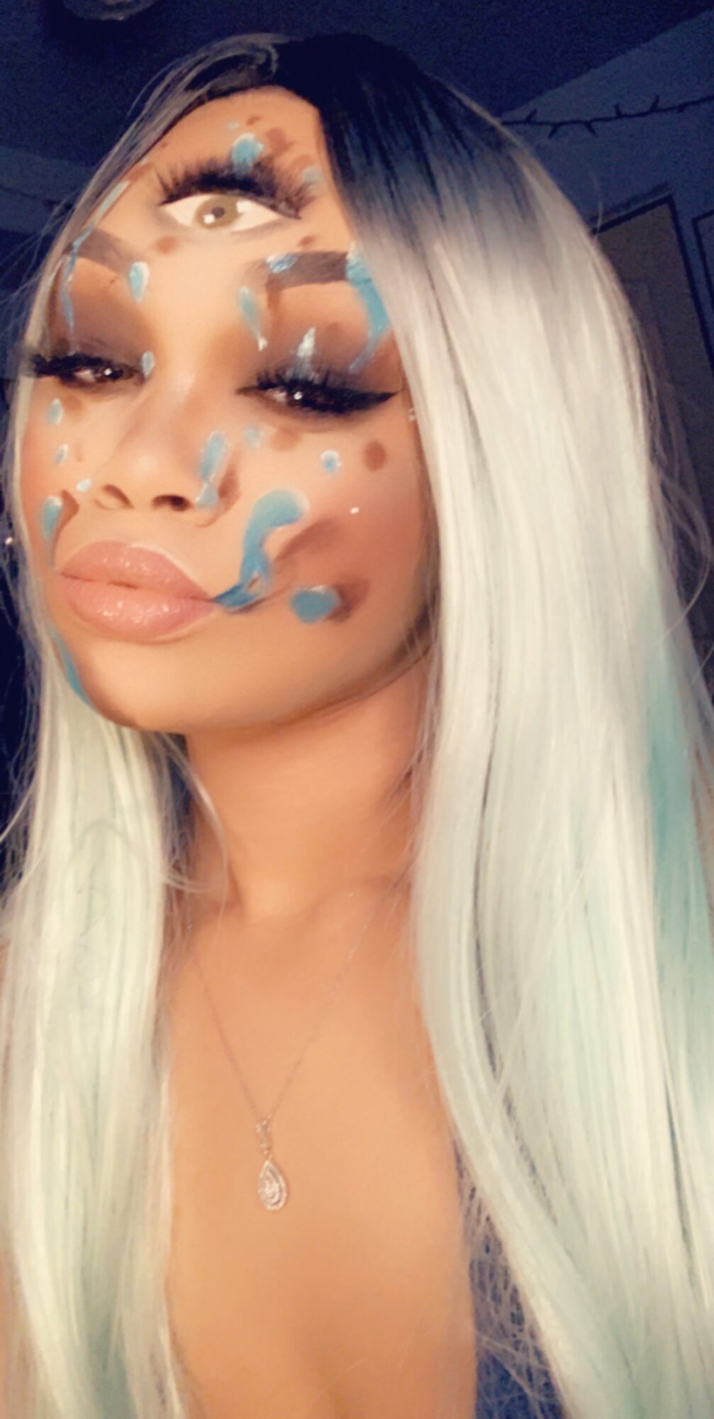 Close-up of a woman with long, two-tone hair, makeup, and blue paint splatters on her face, looking at the camera.