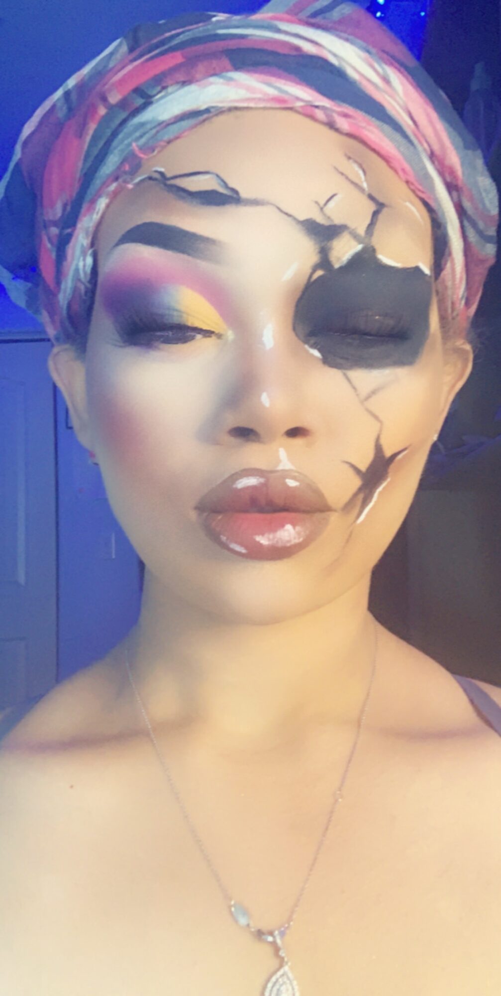Woman with colorful headscarf and makeup resembling a cracked surface, one eye covered with black makeup, lips slightly parted.