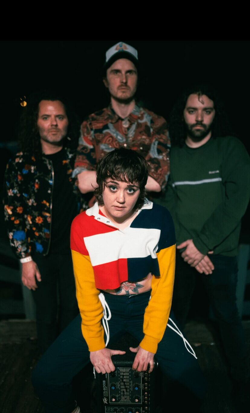 Four people standing outdoors at night, one in front wearing a colorful sweater, three behind in casual clothing.