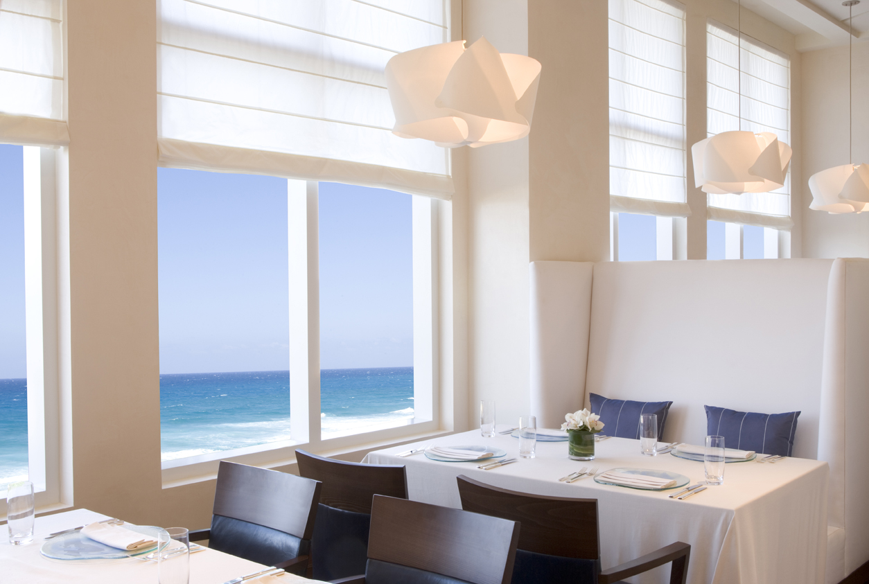 Dining area with large windows showing ocean view, white walls, modern hanging lights, and a table with place settings.