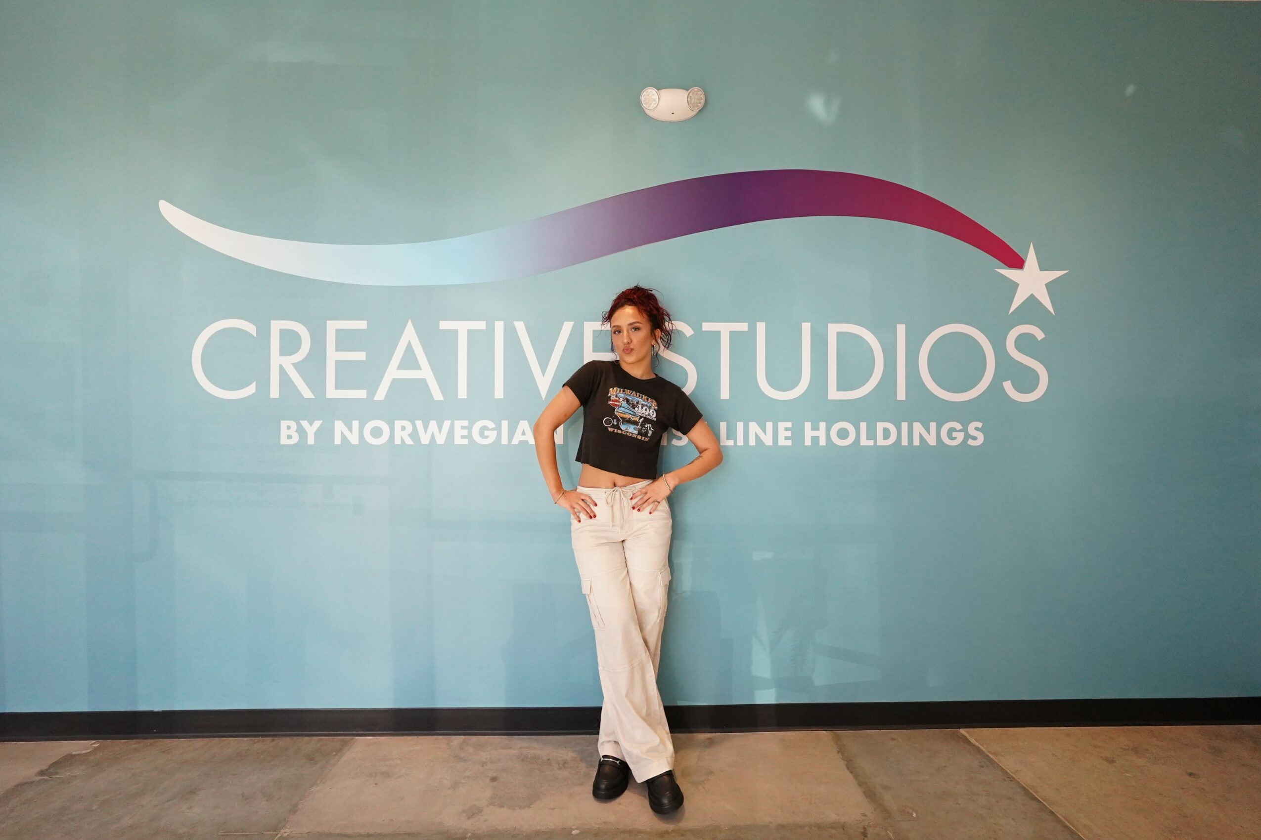 Woman standing in front of a blue wall with a logo and text, hands on hips, smiling, wearing a black shirt and white pants.