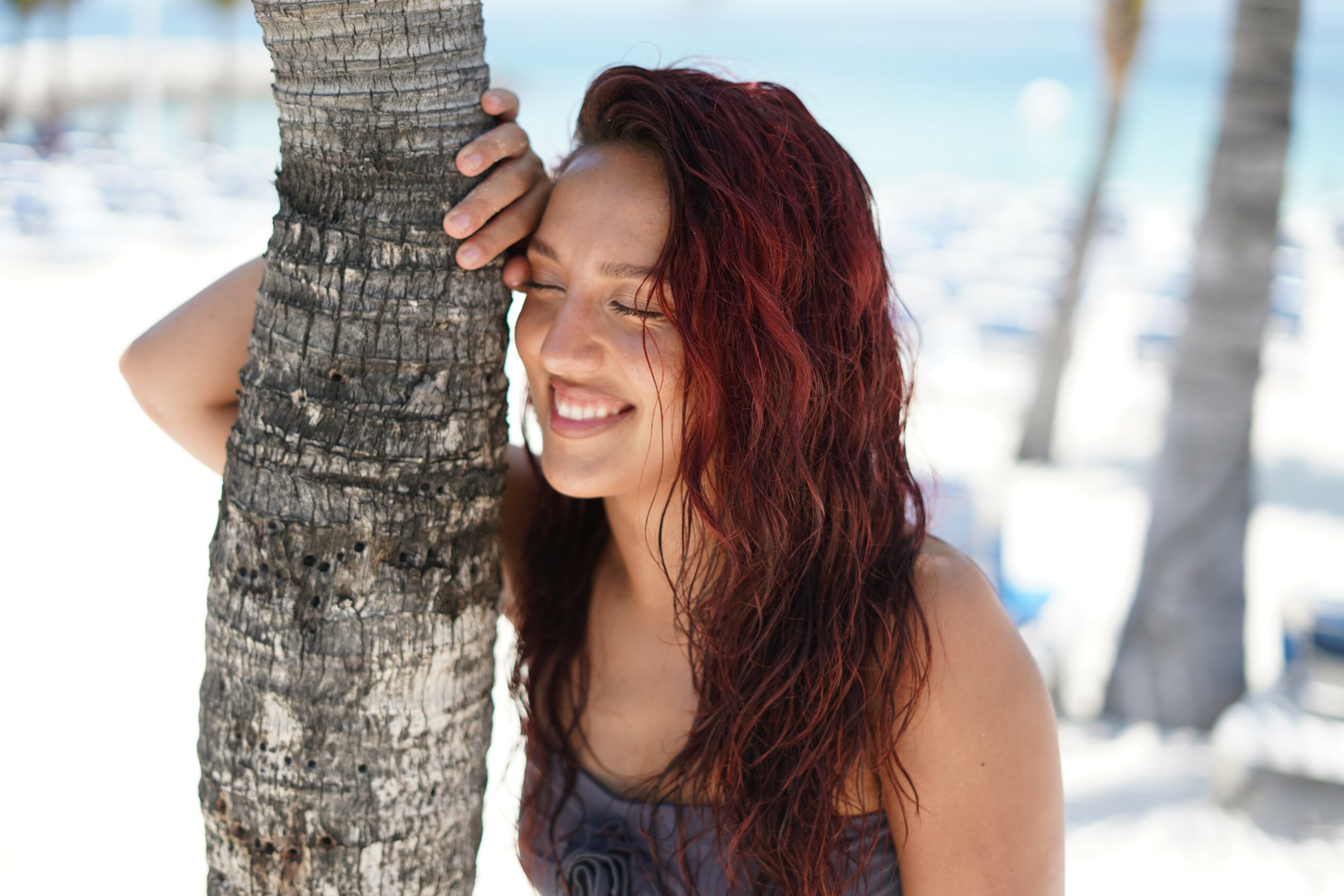 Woman with red hair leaning against a palm tree, smiling with eyes closed, in a beach setting.