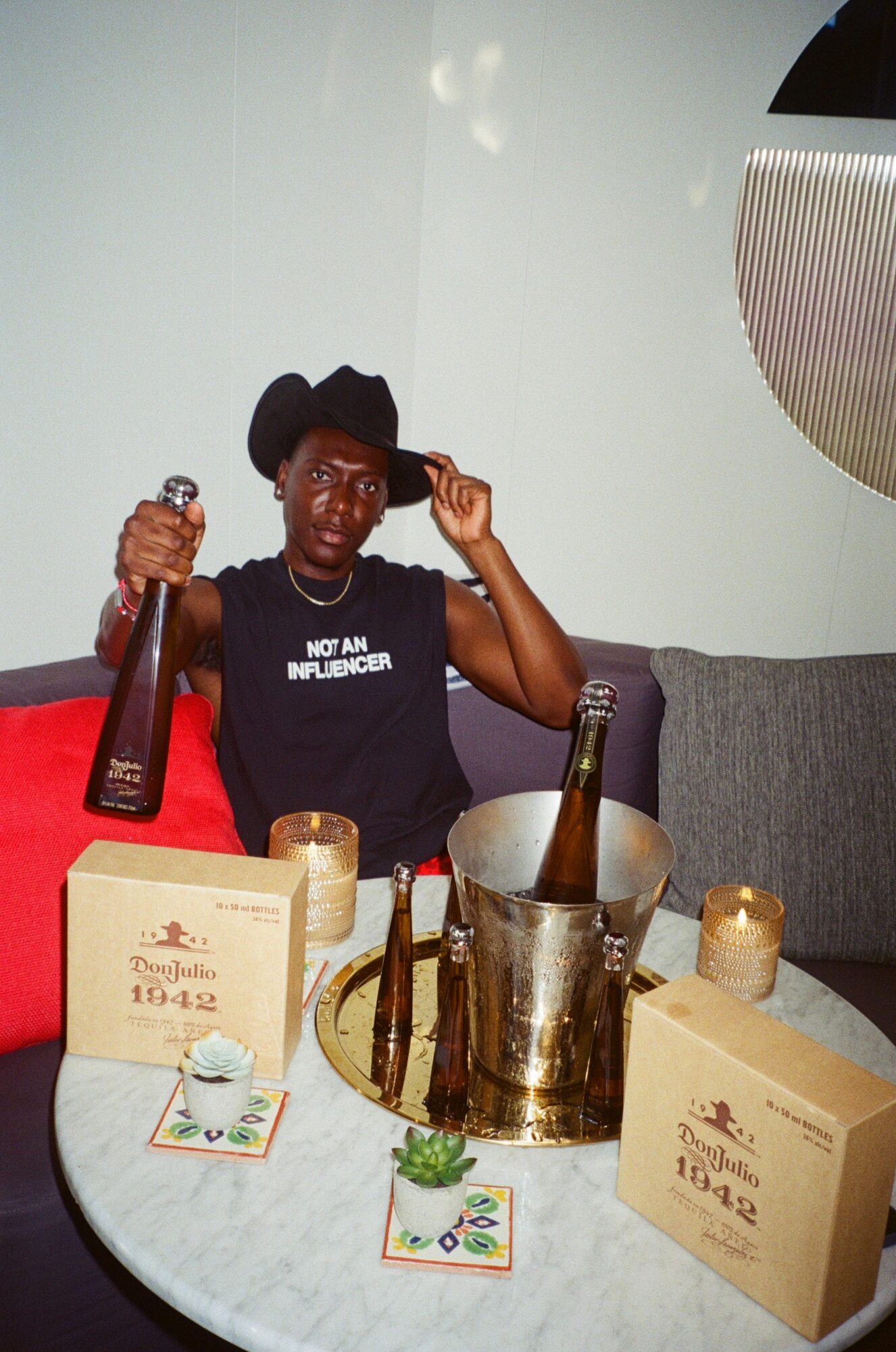 Person wearing a black hat and shirt, sitting at a table with bottles, candles, and boxes, holding a bottle and smiling.