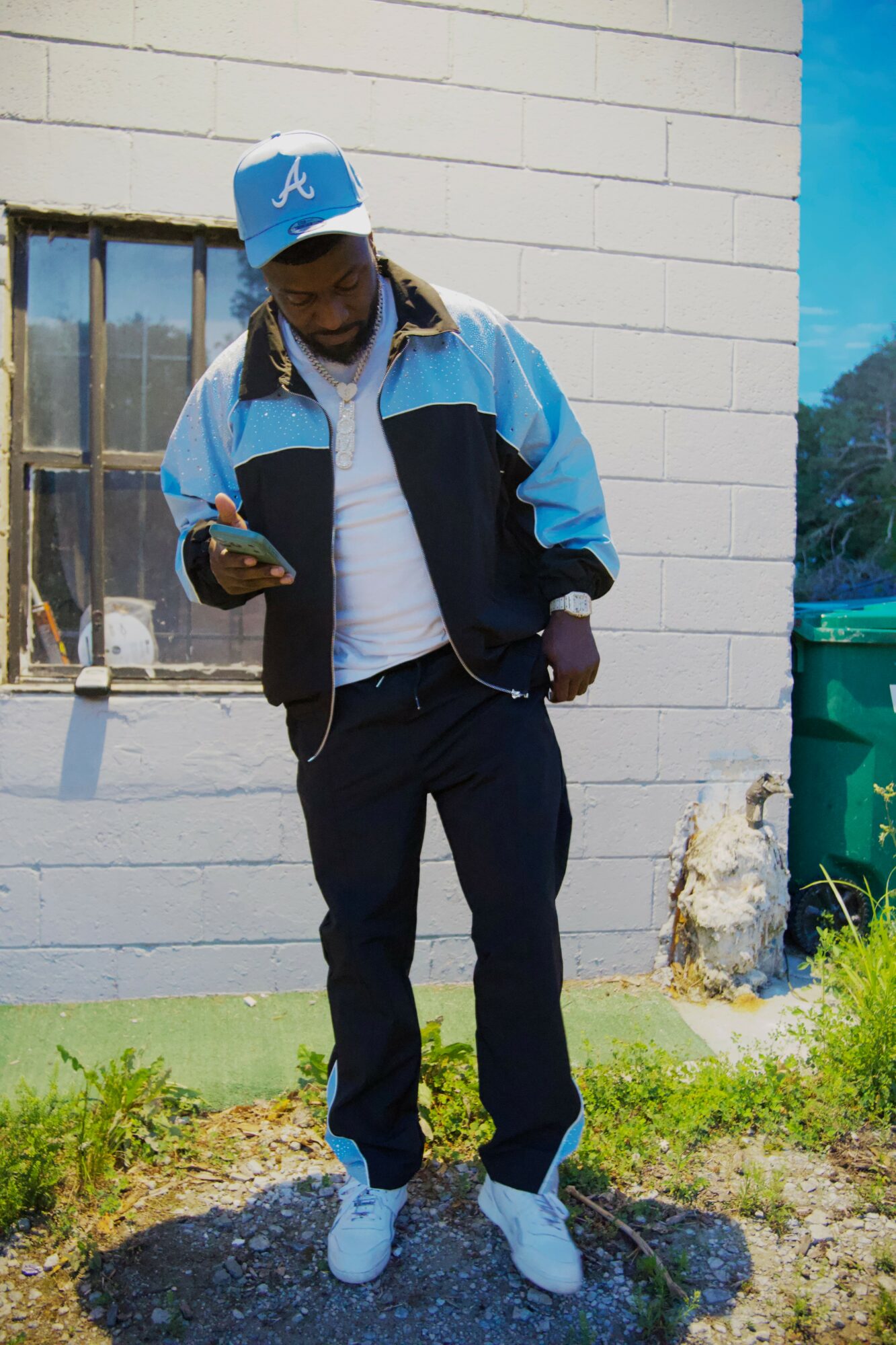 Young person in a blue and black jacket, white sneakers, and a blue cap, standing outdoors and looking at a phone.