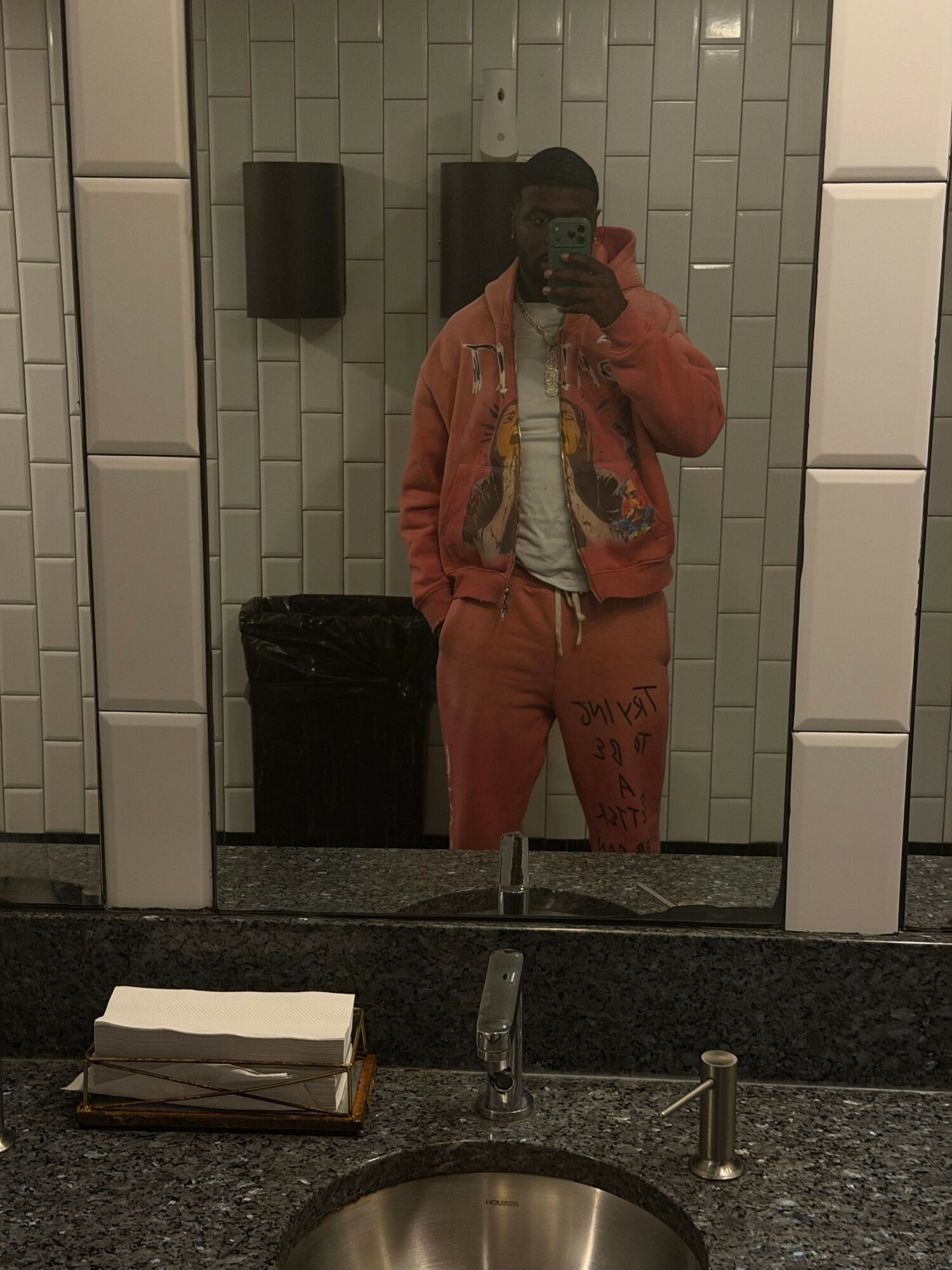 Person standing in front of a mirror in a bathroom with white tiled walls, wearing an orange jacket and pants, holding a phone.