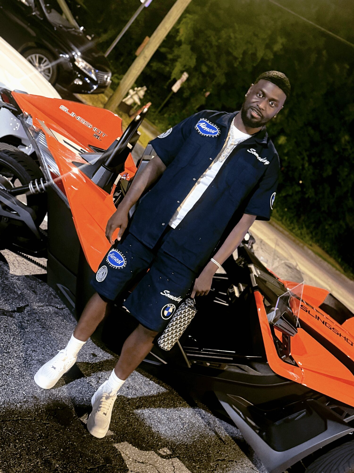 Young man standing next to an orange sports car outdoors at night, wearing a dark vest, shorts, and white shoes.