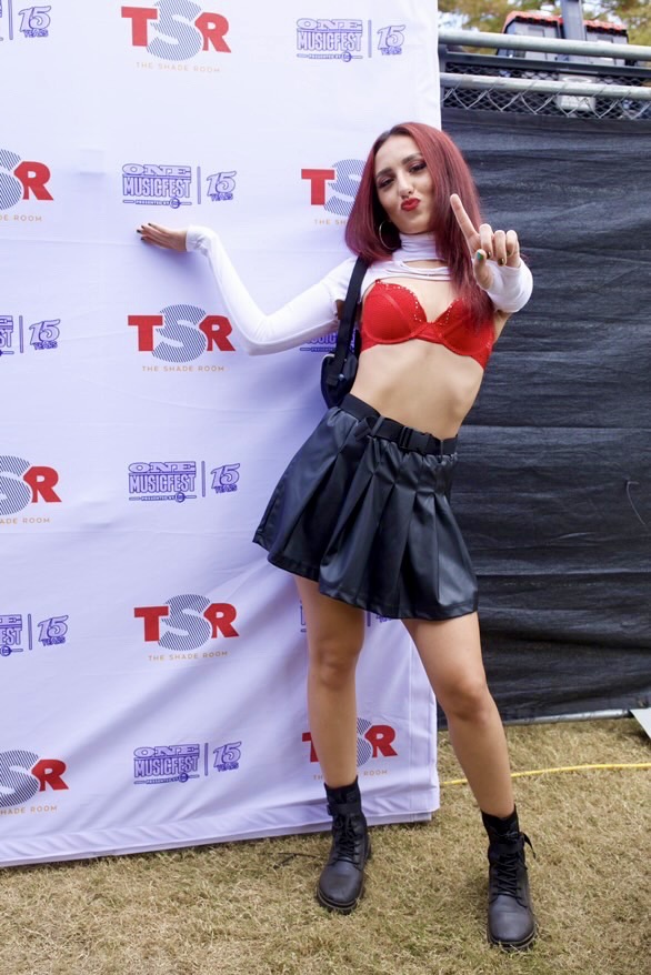 Young woman with red hair in a red and white crop top, black skirt, and boots, posing outdoors against a backdrop.