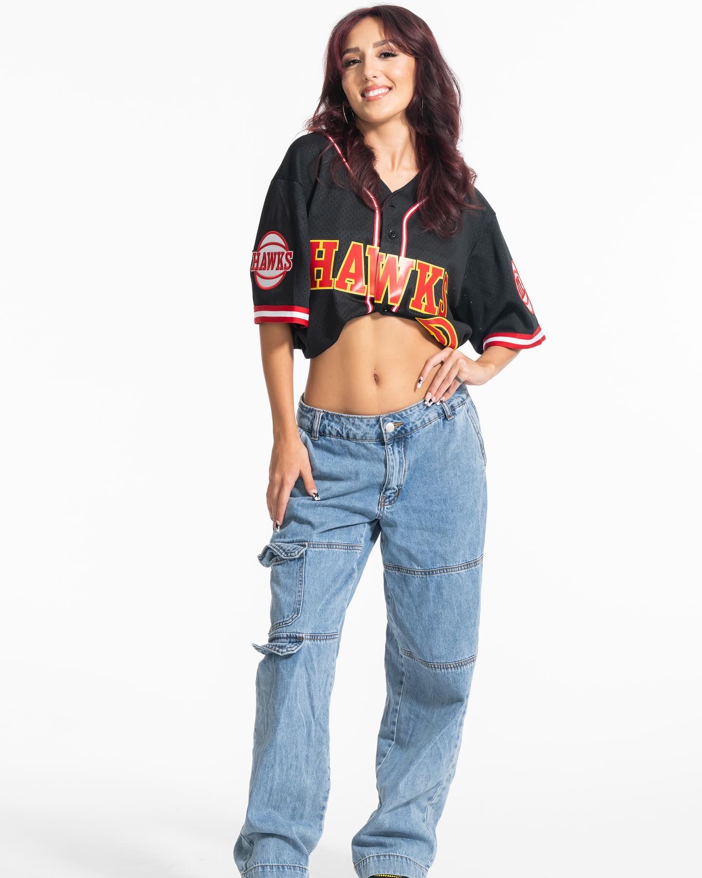 Young woman with dark hair smiling, wearing a black sports jersey and loose jeans, standing against a white background.