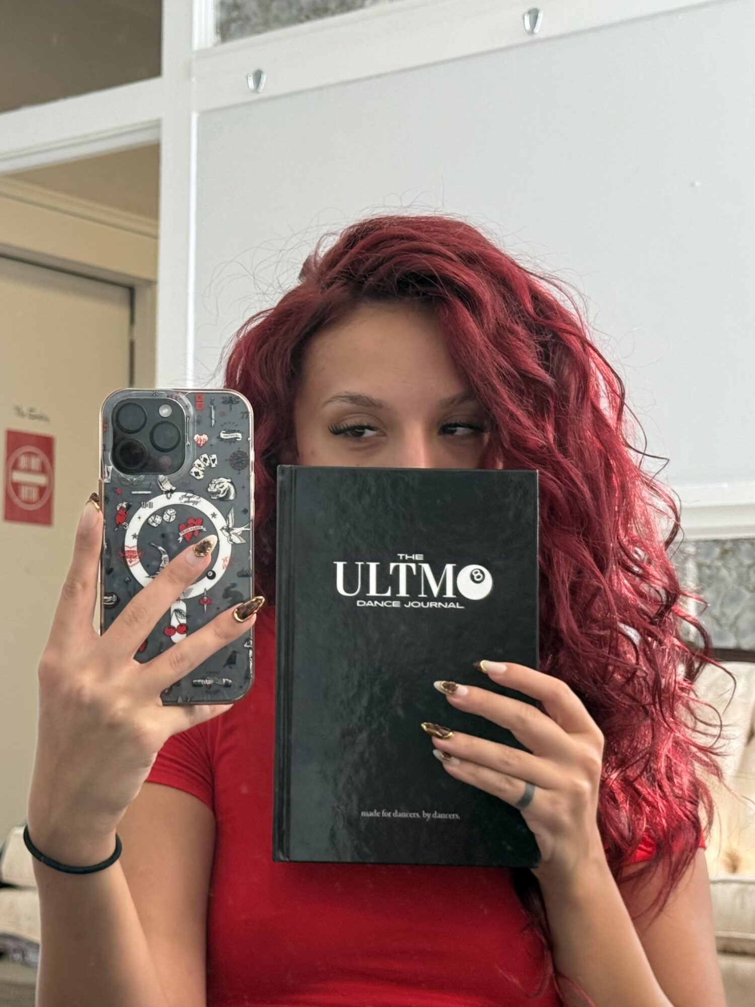 Person with curly red hair holding a black book titled 'ULTMO' and taking a selfie in a mirror, wearing a red shirt.