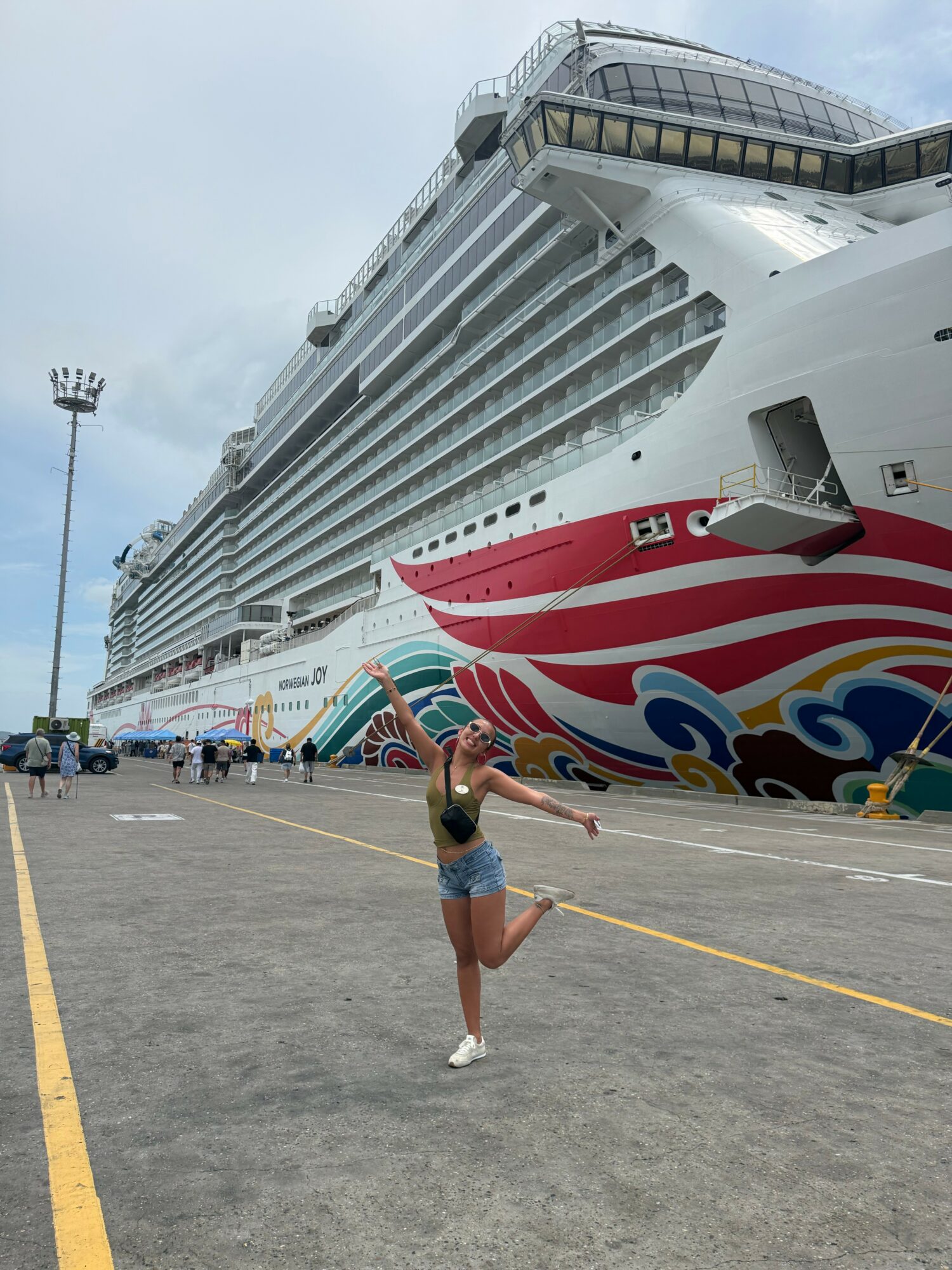 Person standing on dock near large cruise ship, holding a phone, with cloudy sky in background.