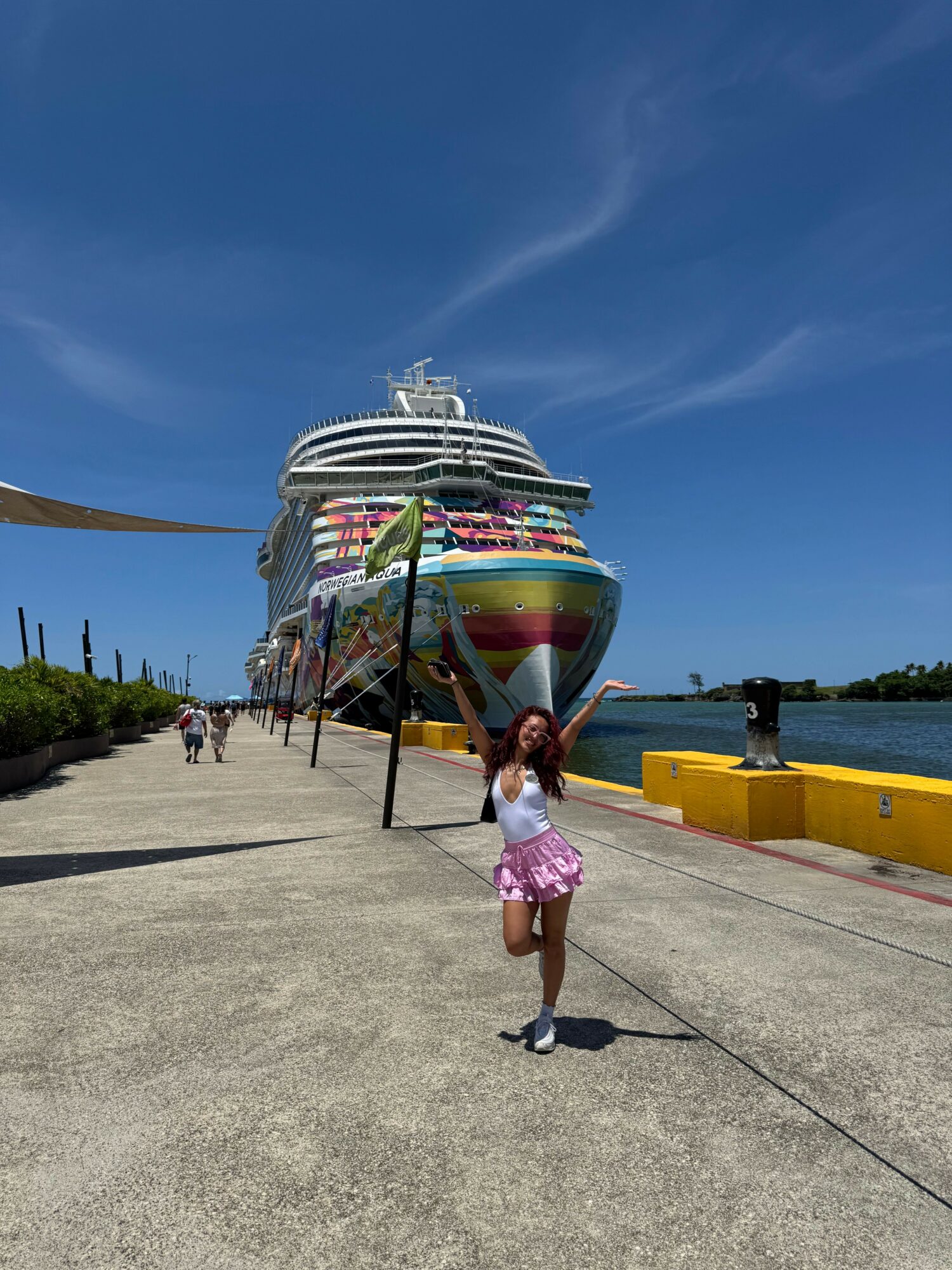 Child in pink shorts and black shoes stands on concrete near a colorful boat, with a blue sky and water in background.