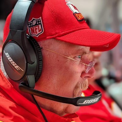 Man wearing a red cap and Bose headset, looking to the right, with glasses and a beard, outdoors.