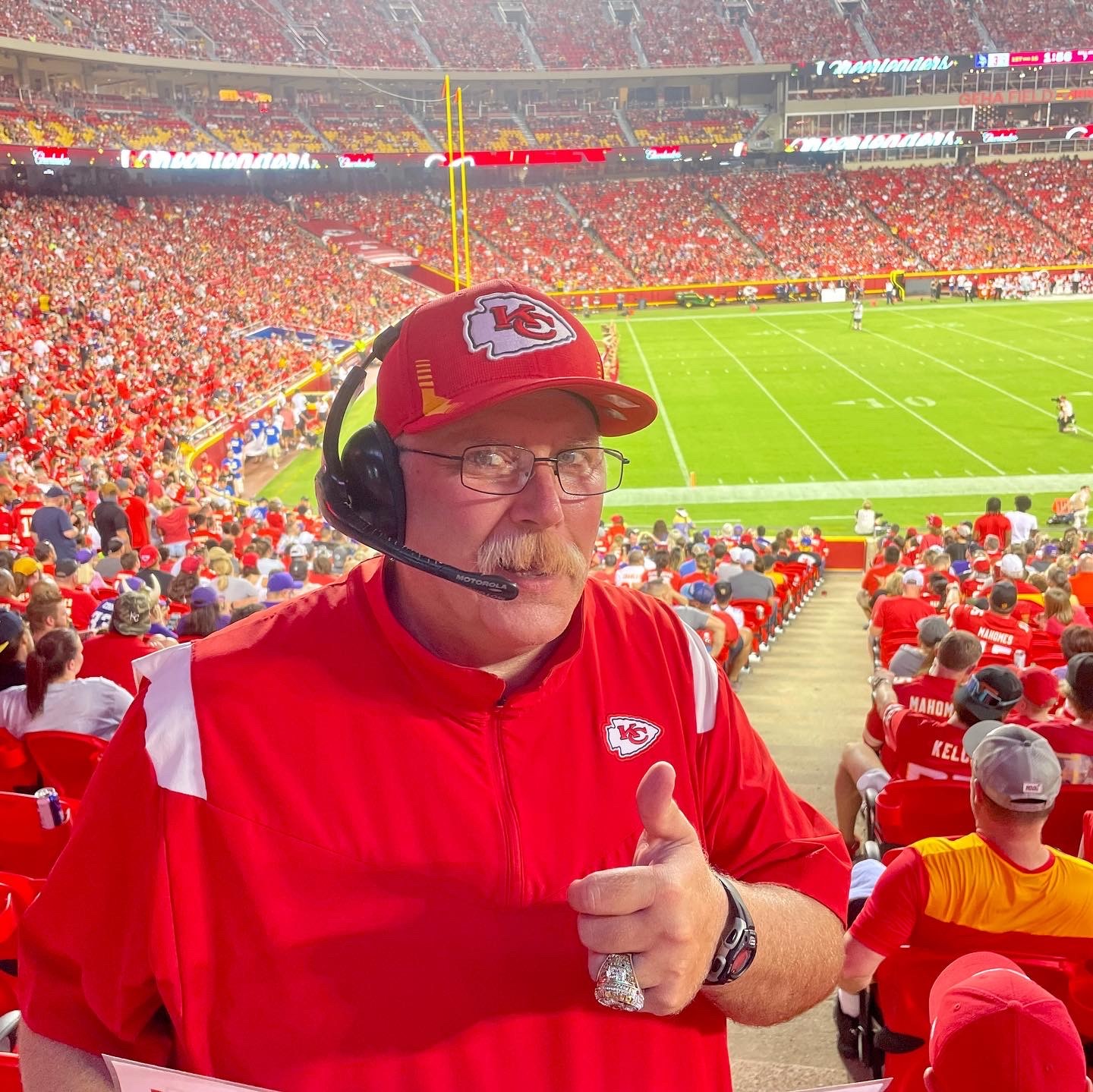 Man wearing a Kansas City Chiefs cap and red jacket, giving a thumbs-up at a football stadium.