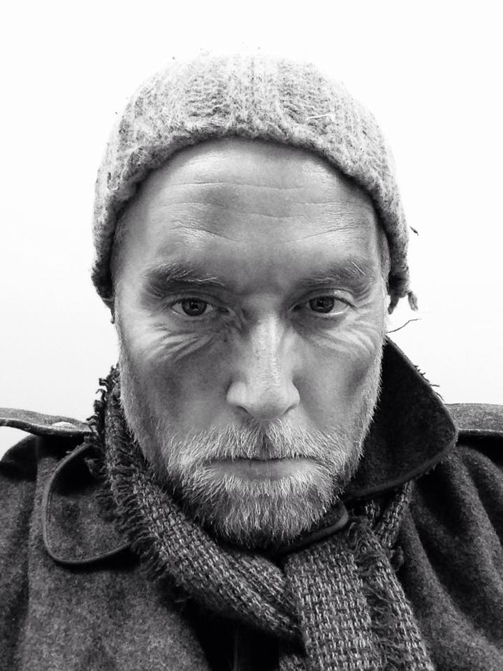 Portrait of a man wearing a knit hat and scarf, with a serious expression, in black and white.
