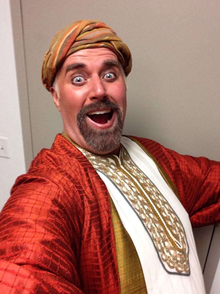 Person with a turban and colorful clothing smiling widely, taking a selfie indoors against a plain wall.