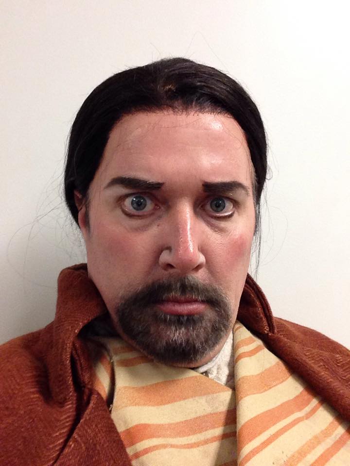 Person with dark hair, beard, and mustache, wearing a striped shirt and brown jacket, looking directly at the camera.