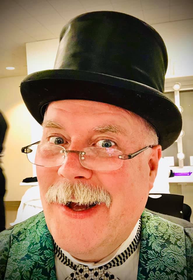 Man with glasses, mustache, wearing a top hat and patterned jacket, smiling indoors.