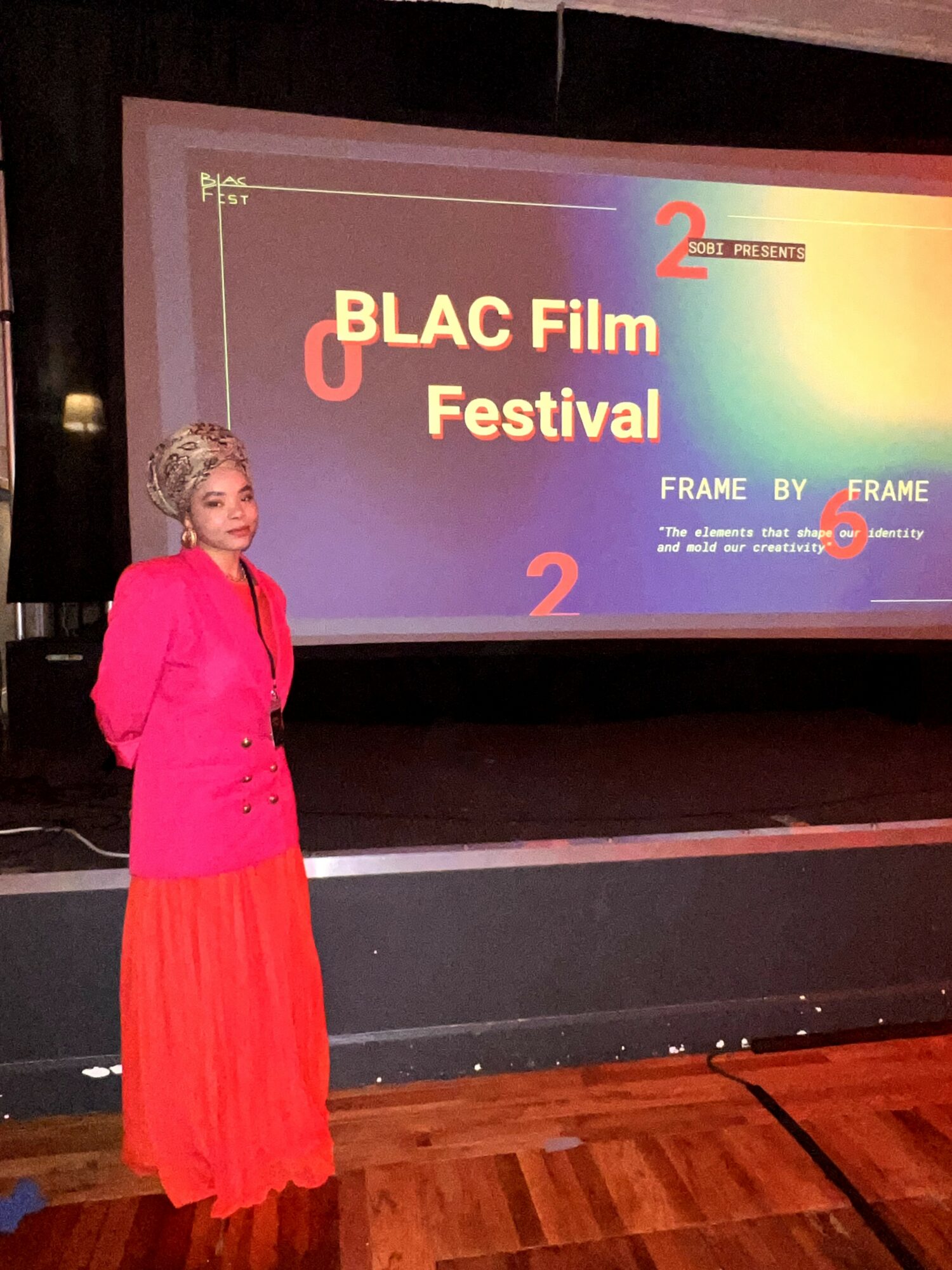 Woman in pink top and red skirt standing in front of a screen displaying 'BLAC Film Festival' with colorful text and graphics.