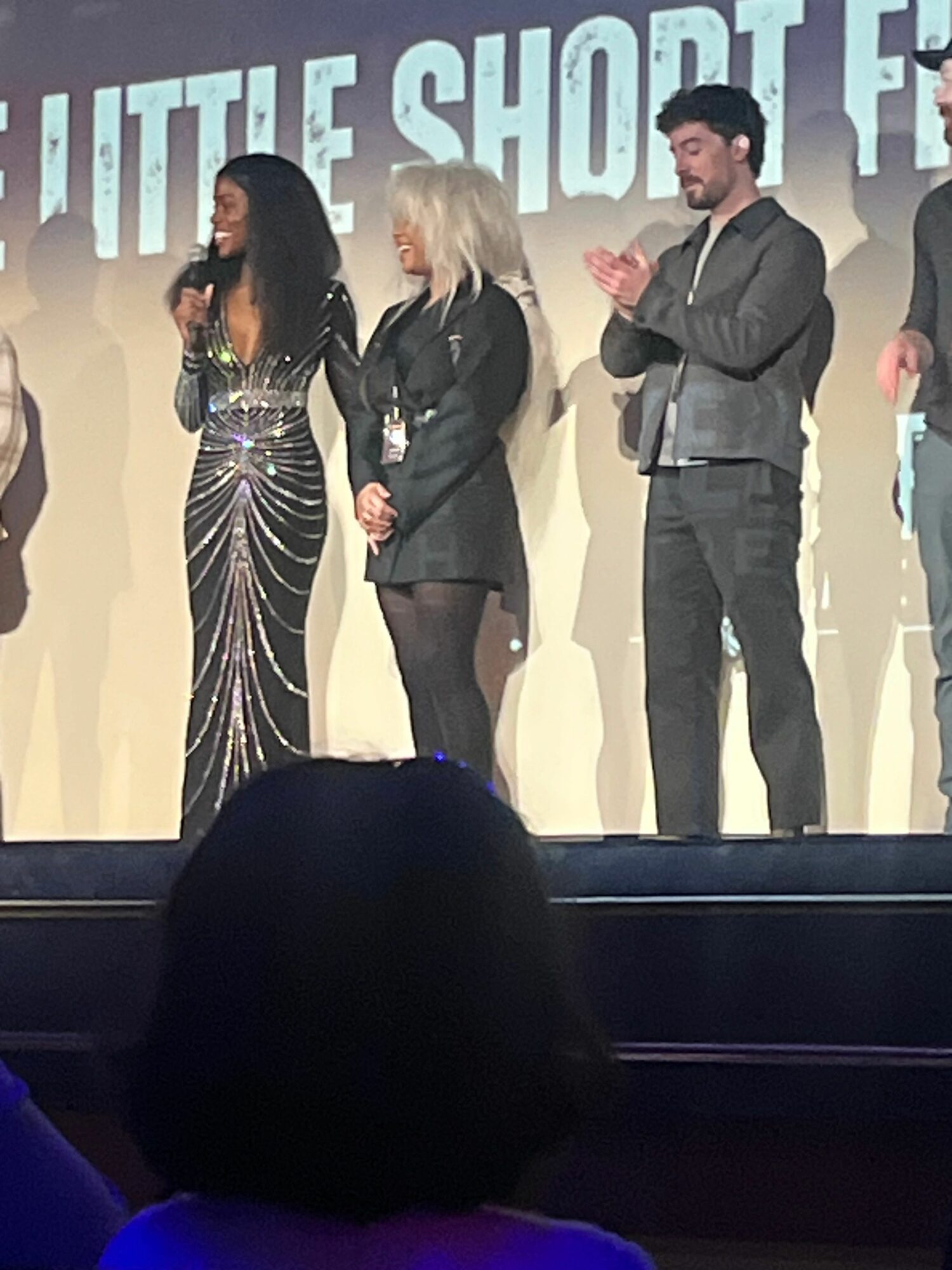 Three people on stage, two women and one man, with a backdrop and audience in foreground.