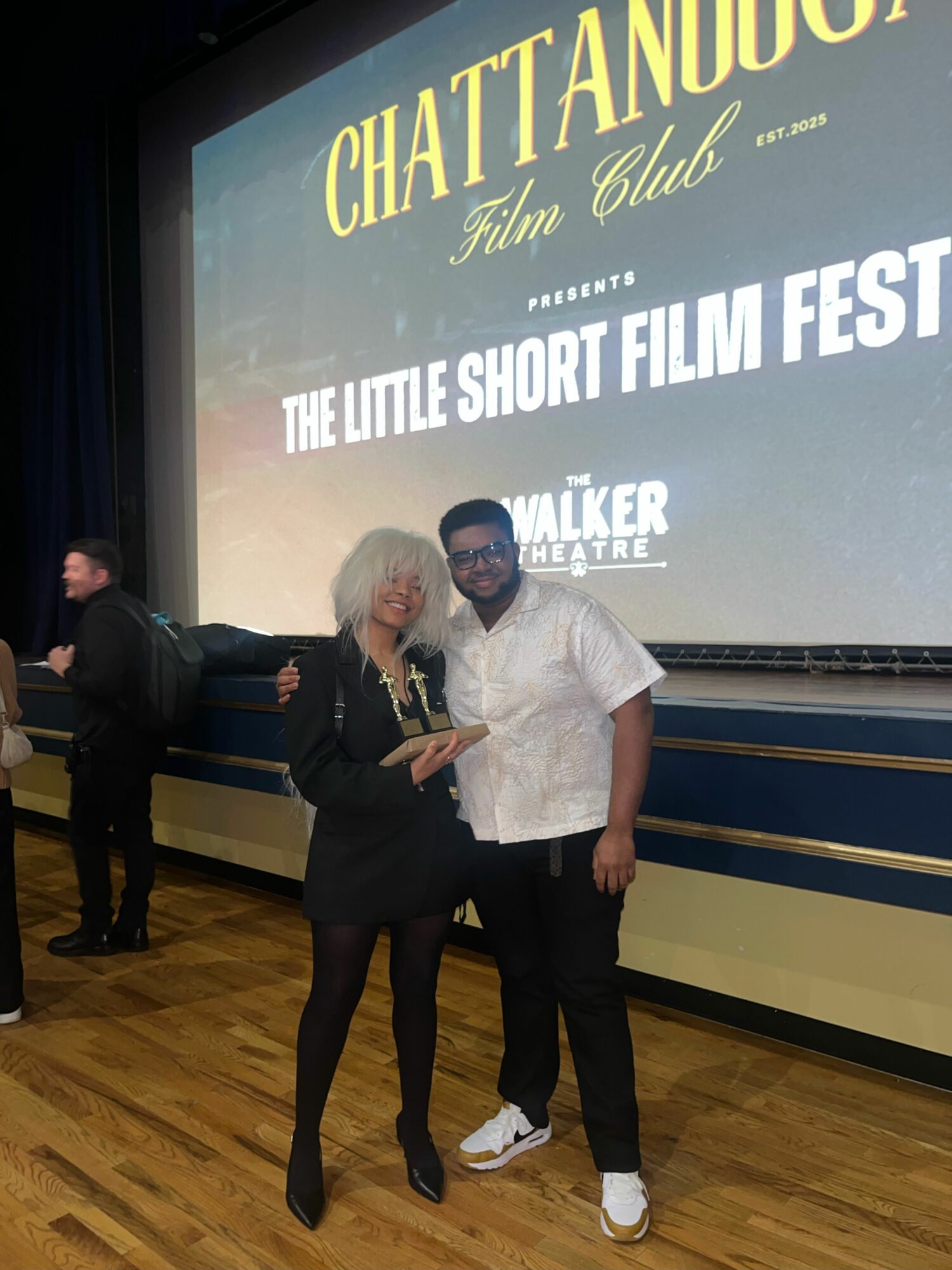 Two people standing in front of a screen at an event, one holding a trophy, with a wooden floor.