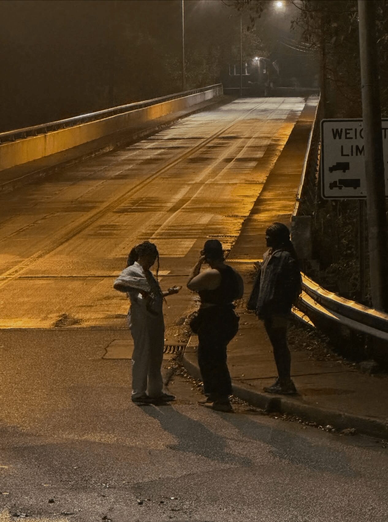 Three people standing on a wet road at night, talking, with a sign and guardrail nearby.