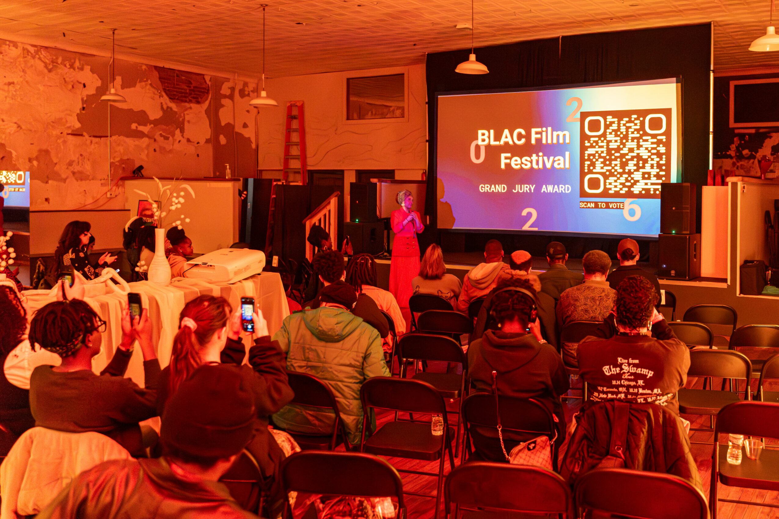 Audience watching a presentation on stage at a film festival, with a large screen displaying a QR code and event details.