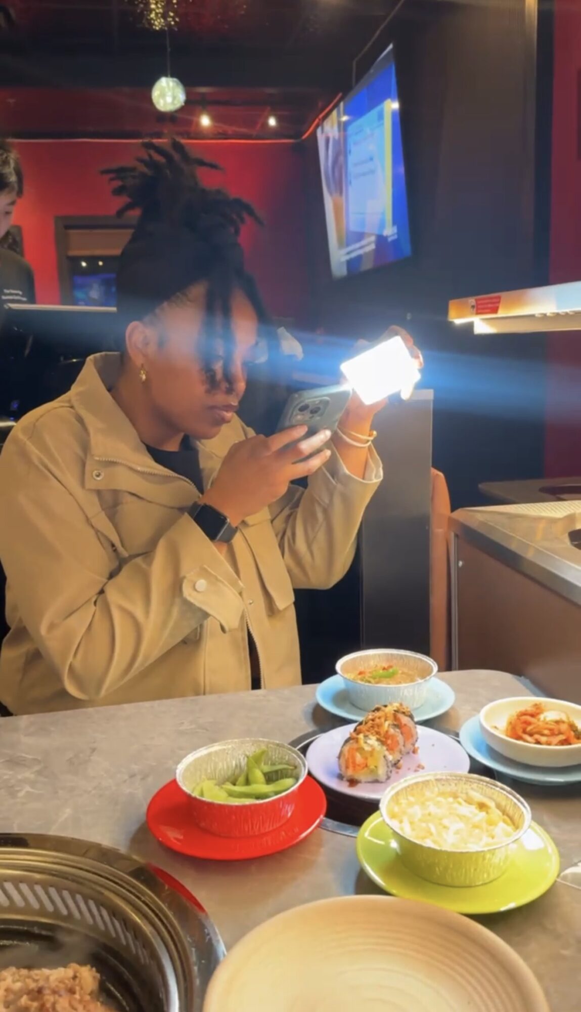 Person with dreadlocks wearing a yellow jacket looking at a phone at a table with various bowls of food.