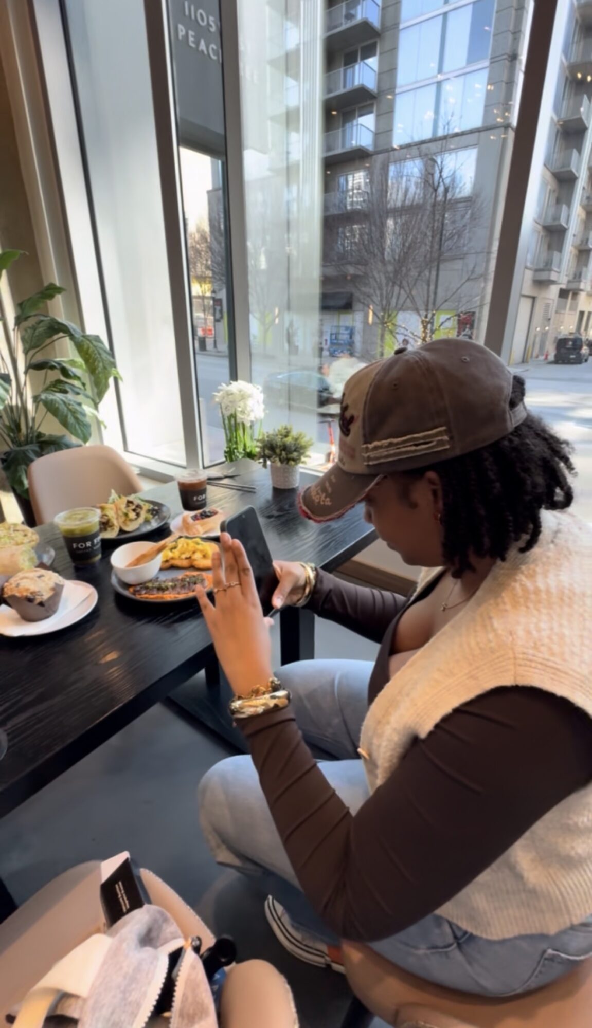 Person with curly hair, wearing a cap and glasses, sitting at a table with food, in a bright cafe with large windows.
