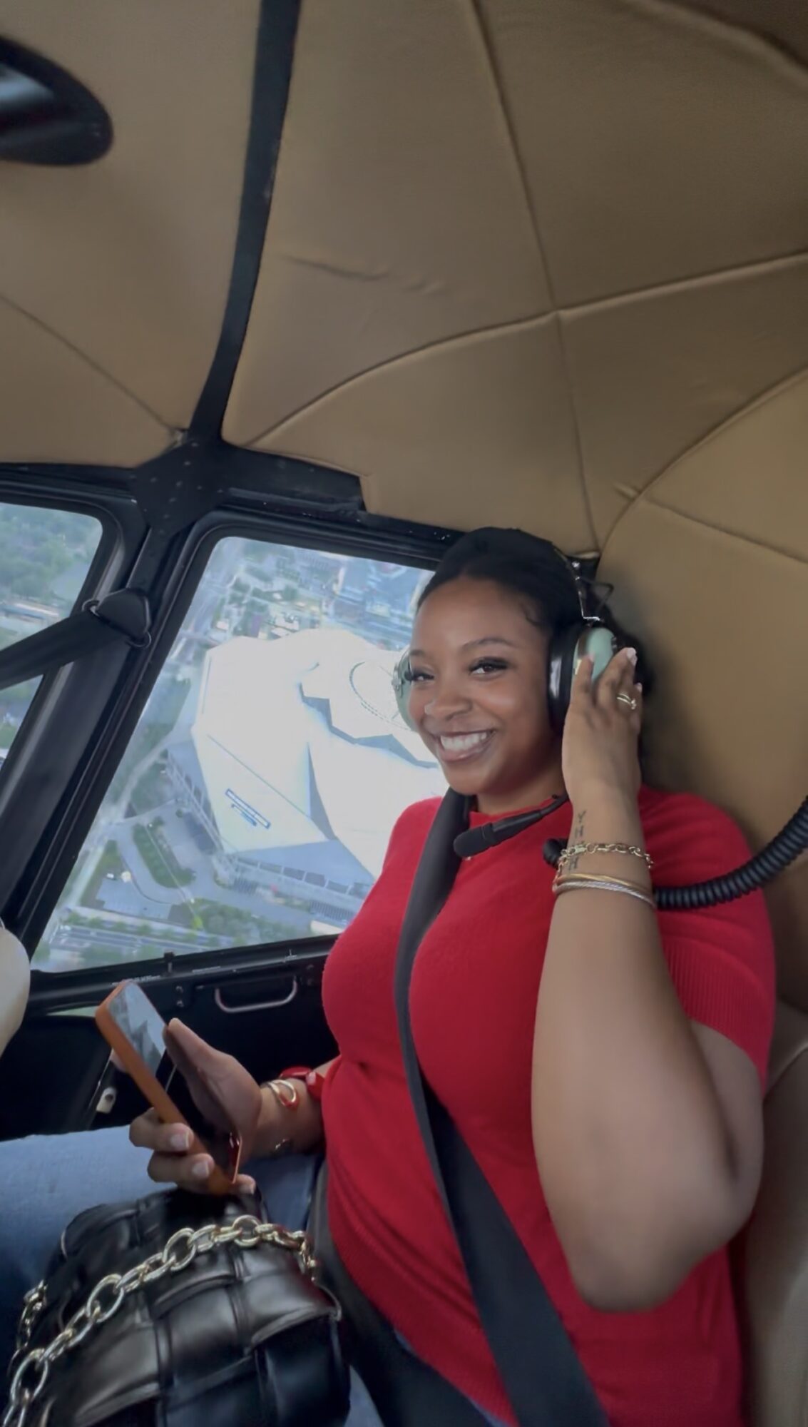 Woman in red shirt smiling, wearing headphones, inside a vehicle, holding a phone, with a window showing a road and trees.