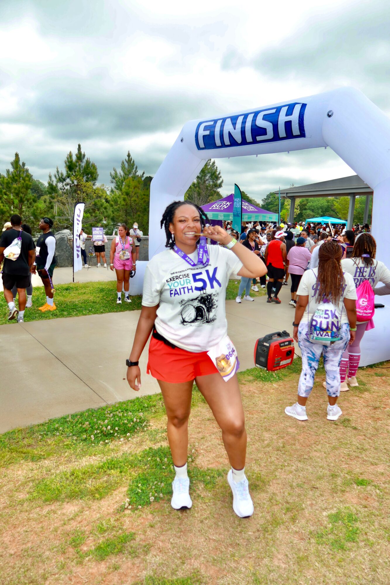 Woman in athletic clothing smiling and drinking water at finish line of race event, crowd and trees in background.